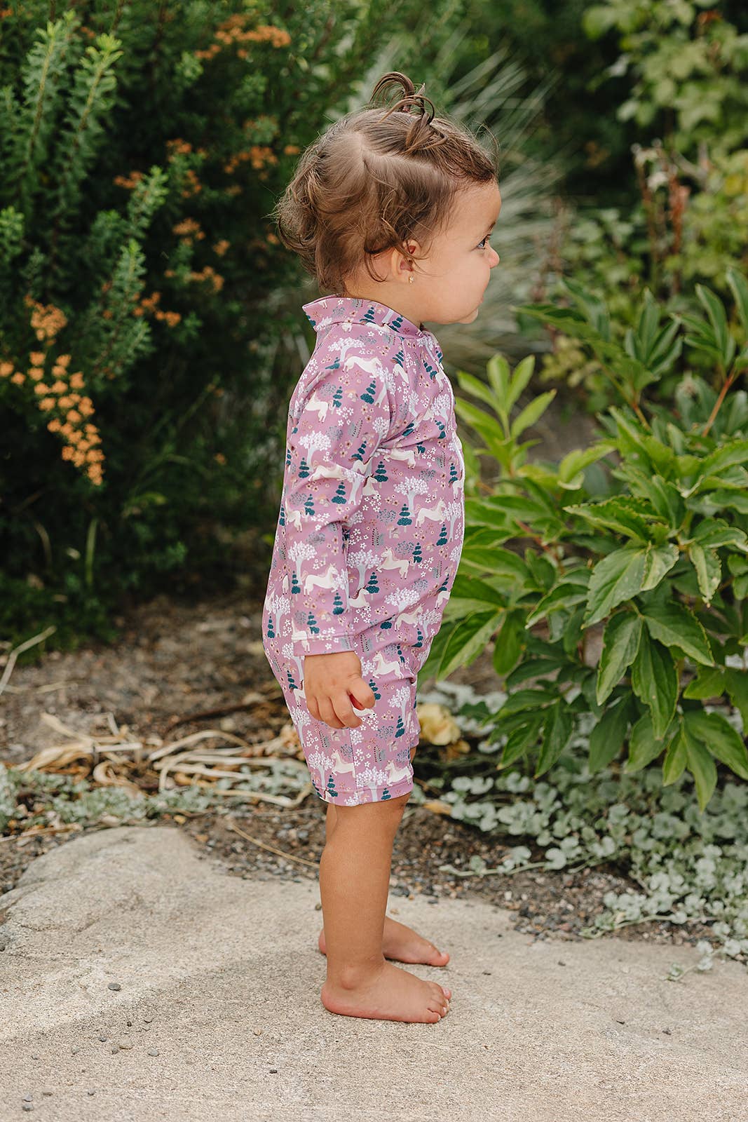 
                  
                    Child wearing a pink unicorn print sunsuit standing outdoors with greenery in the background
                  
                