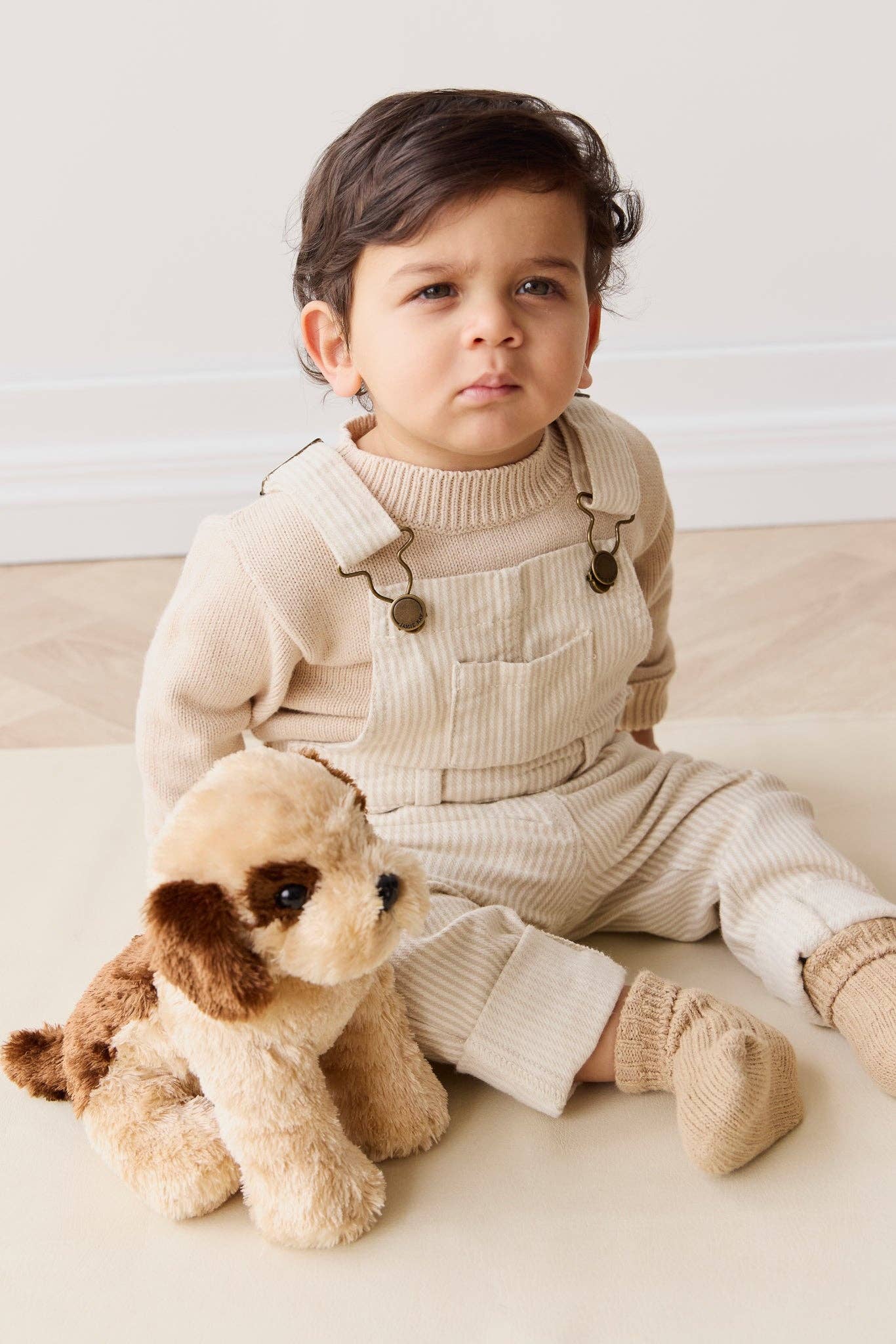
                  
                    Child wearing a beige outfit with suspenders sitting next to a plush toy  dog on a light background.
                  
                