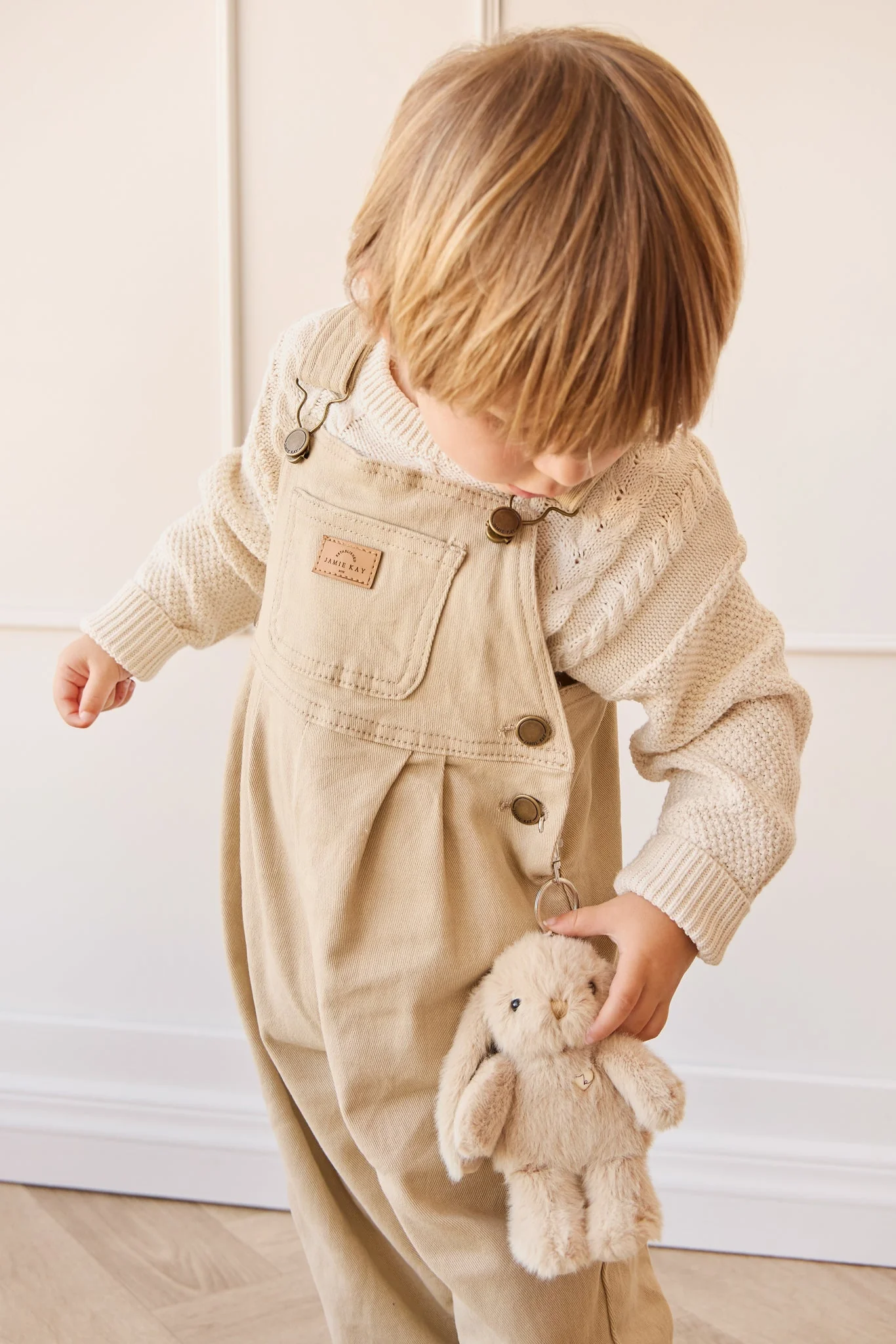 
                  
                    Child wearing beige overalls holding a plush toy against a white wall.
                  
                