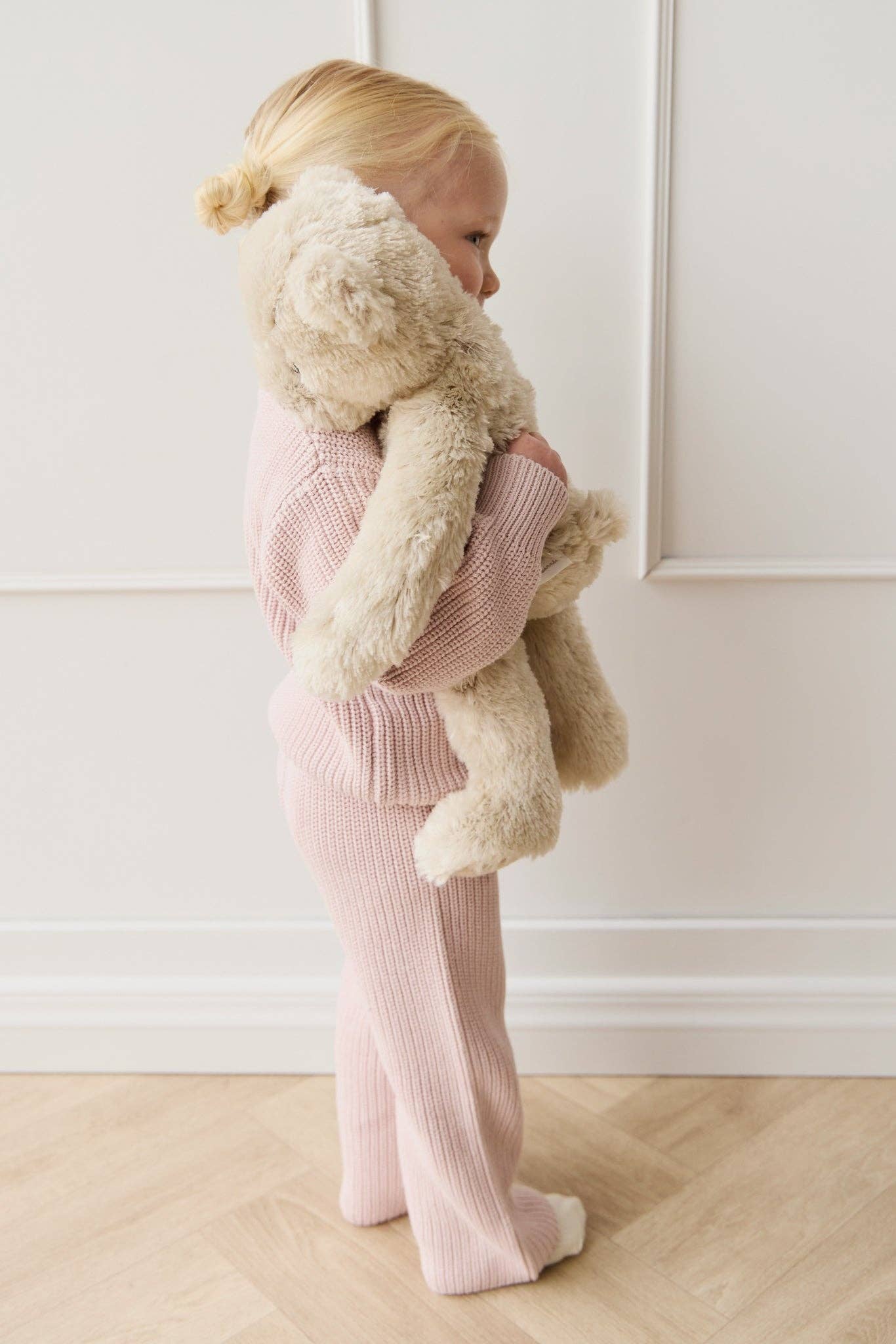 
                  
                    Child in pink outfit holding a large teddy bear against a white wall.
                  
                