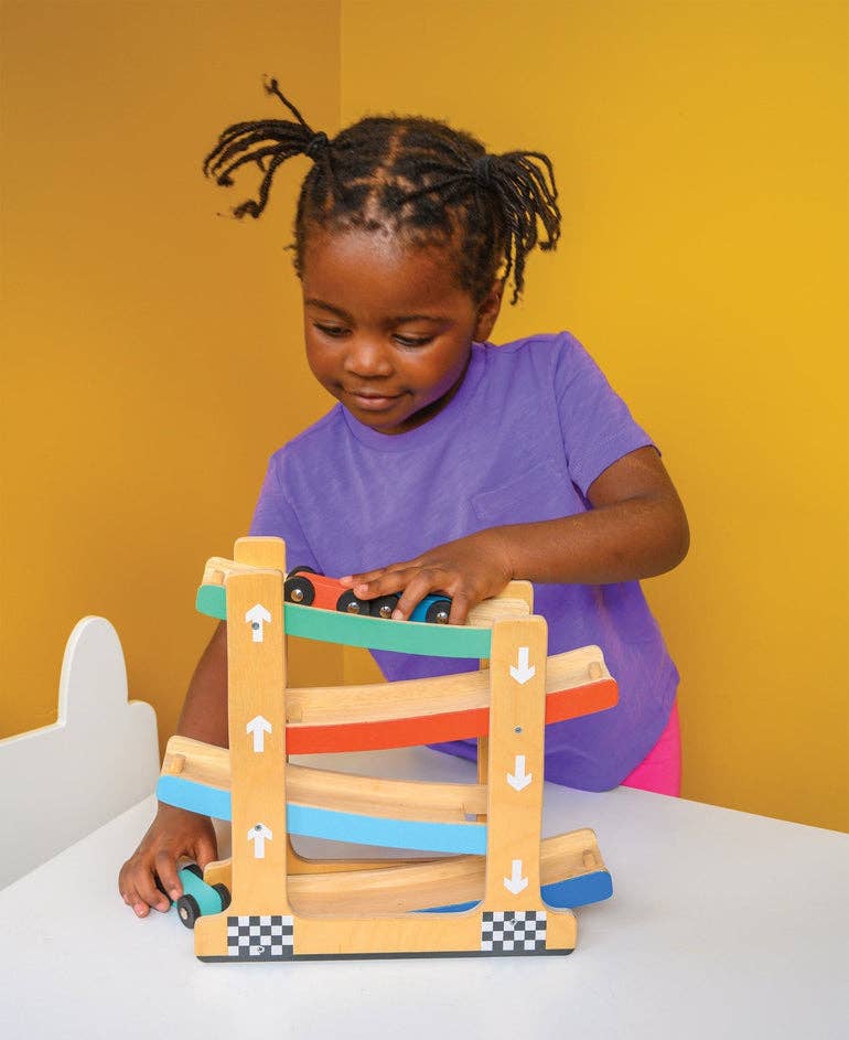 
                  
                    A young child wearing a purple shirt plays with a wooden toy race track on a white table against a solid yellow background.
                  
                