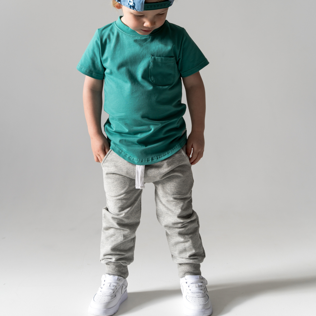 
                  
                    Child wearing a green t-shirt and gray pants on a plain background.
                  
                