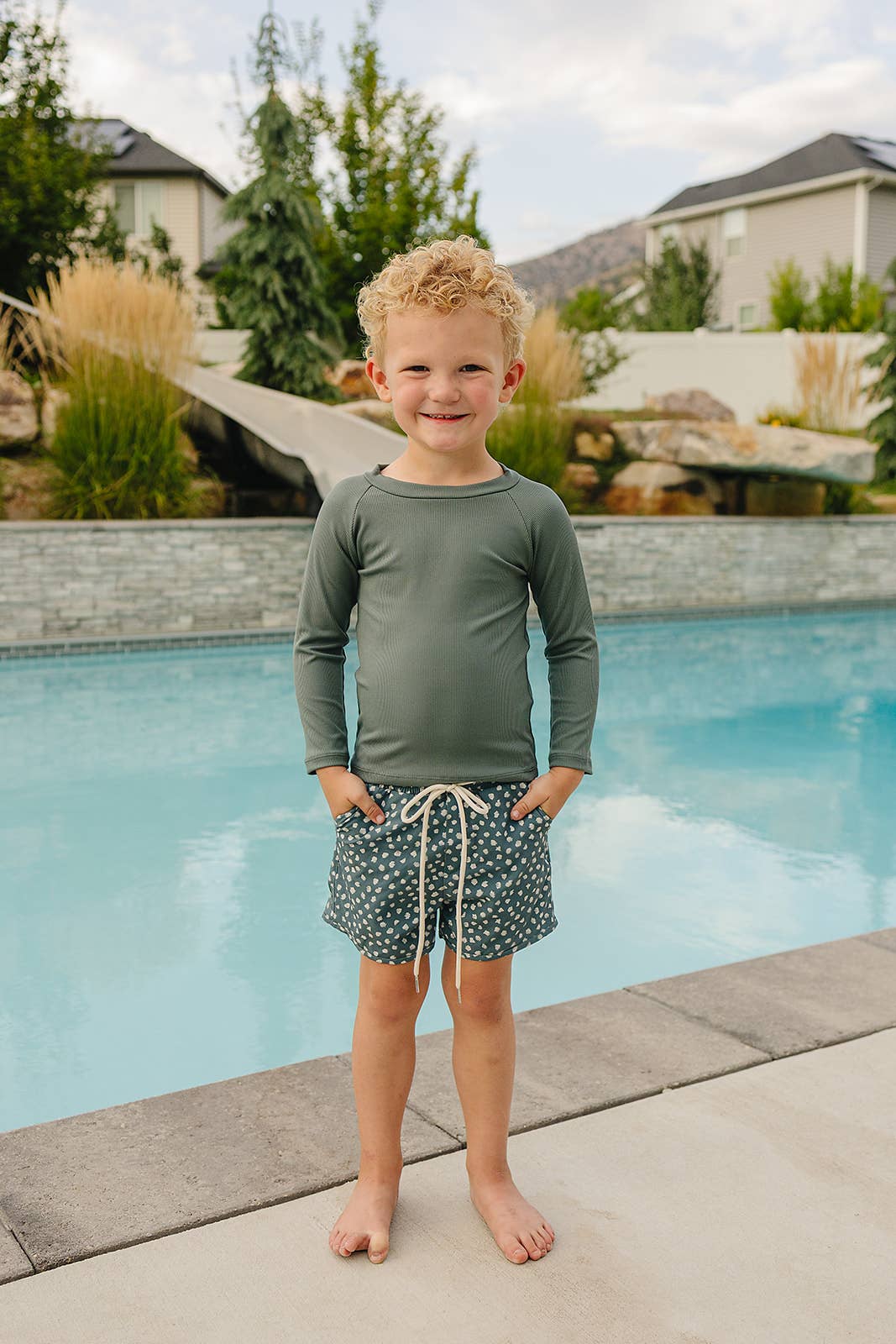 
                  
                    Child standing by a pool wearing a green long-sleeve rash guard shirt and patterned swim shorts.
                  
                