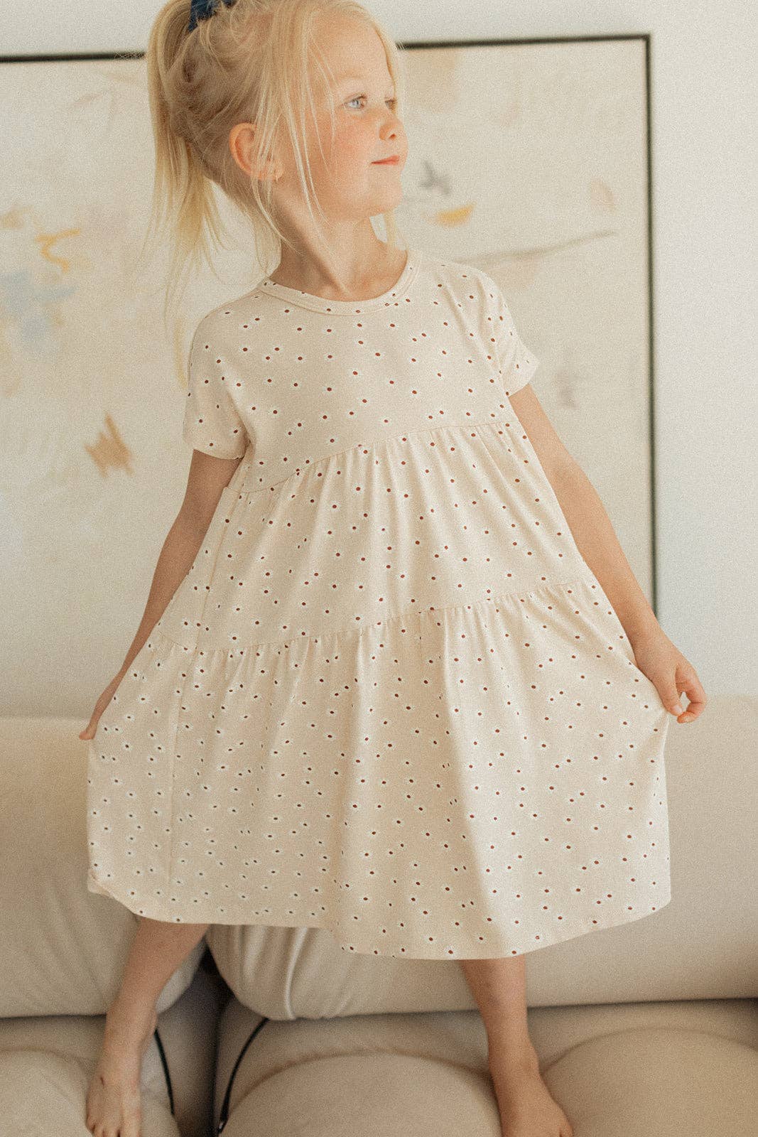 
                  
                    Young girl wearing a white dress with small patterns, standing indoors.
                  
                