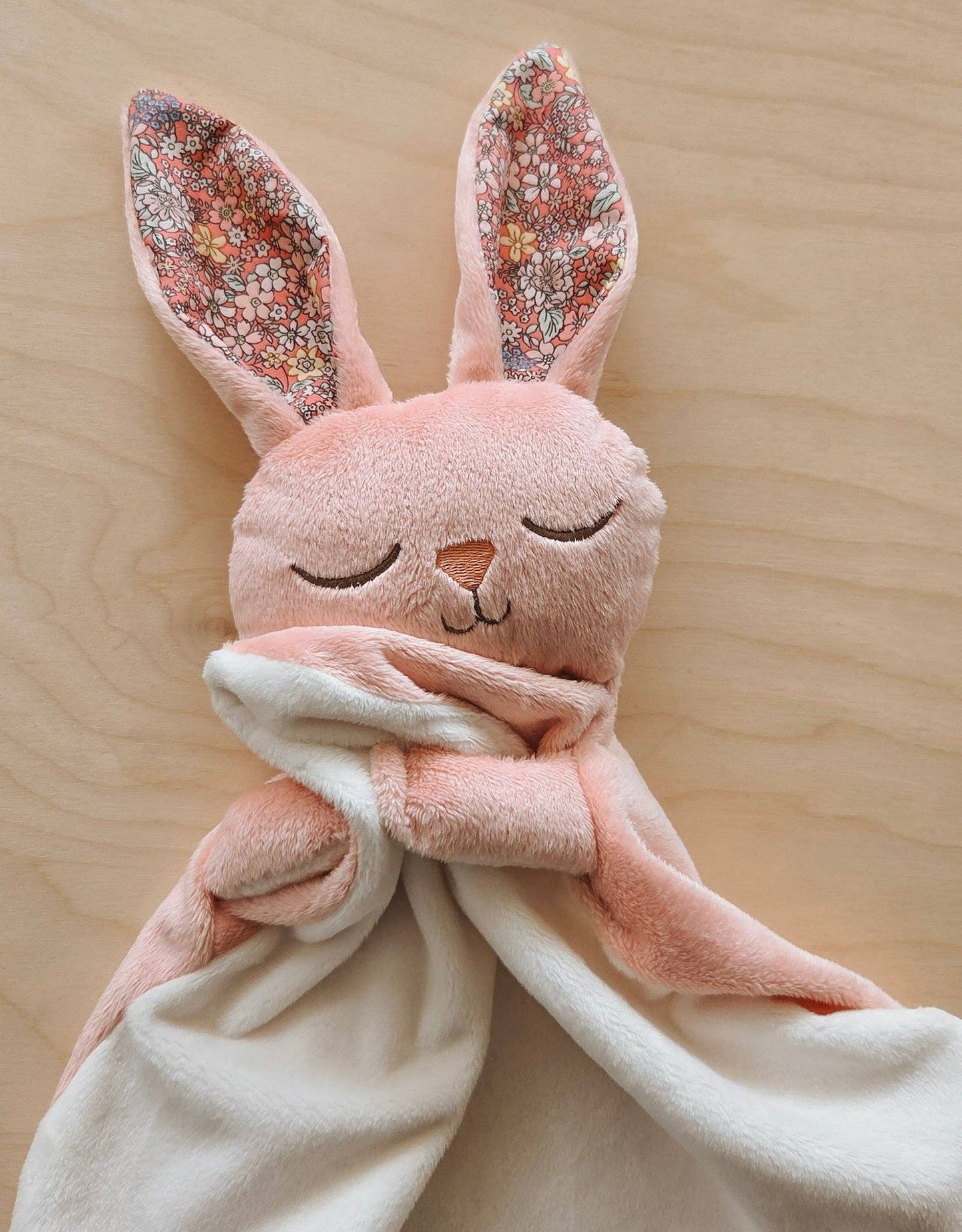 
                  
                    Pink plush bunny with floral ears and a white blanket on a wooden surface.
                  
                
