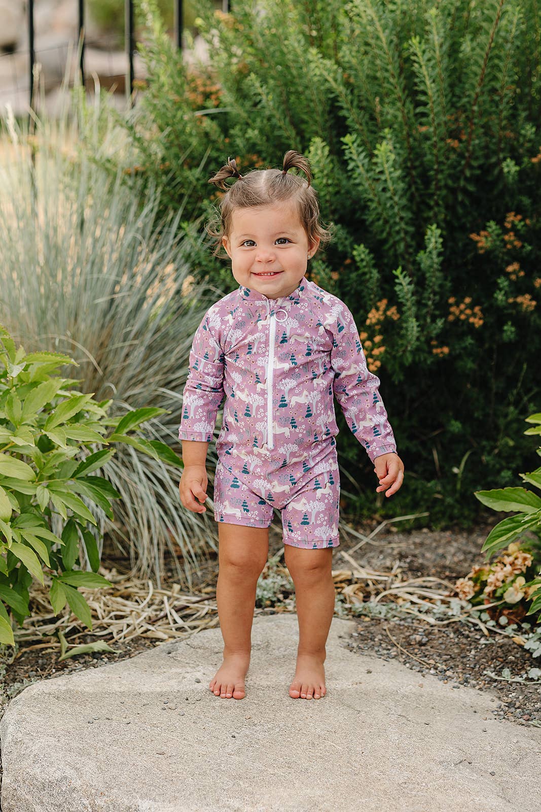 Child wearing a pink and purple unicorn patterned swimsuit standing outdoors among plants.