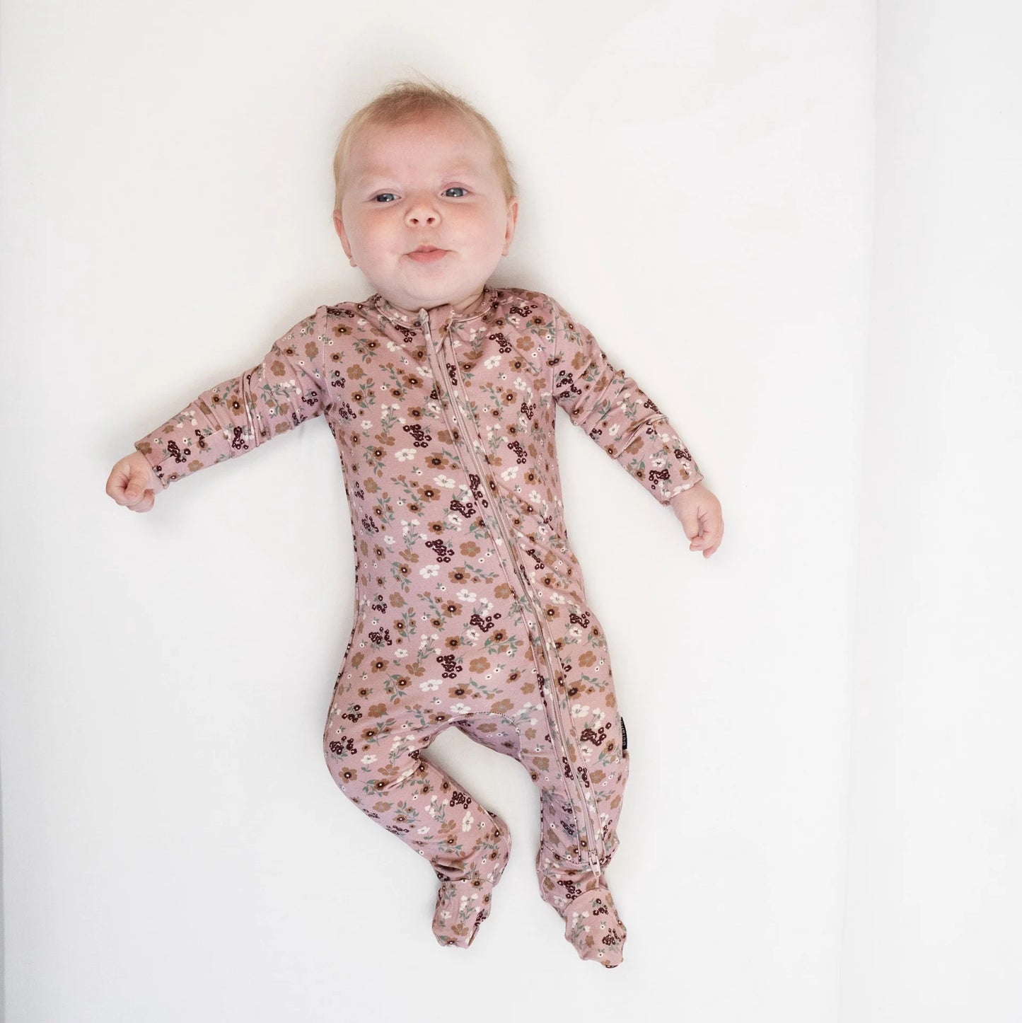
                  
                    A baby lying on their back wearing a long-sleeve, footed zip-up sleeper in a dusty pink and brown floral pattern, set against a plain white background.
                  
                