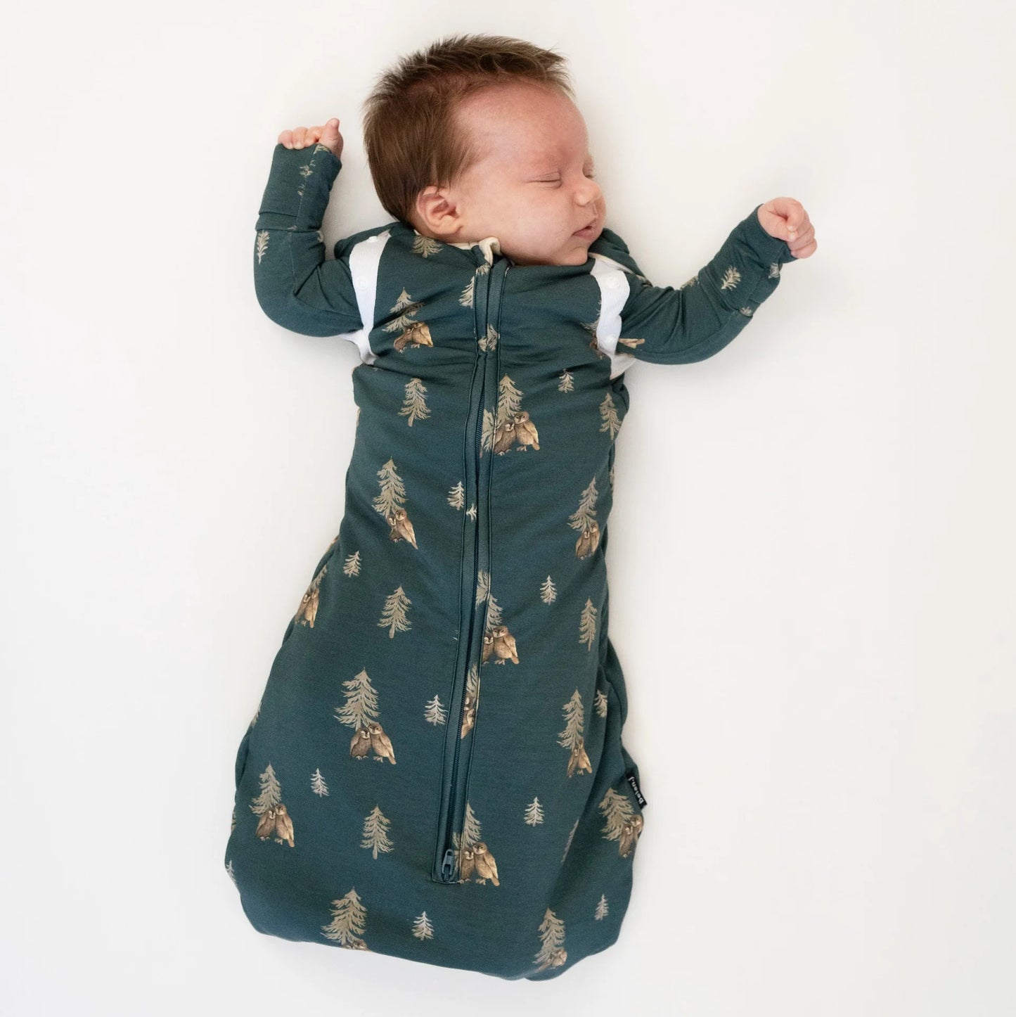
                  
                    A sleeping newborn baby wearing a dark teal green zippered sleep sack with a gold pine tree and owl pattern, lying on a white background.
                  
                
