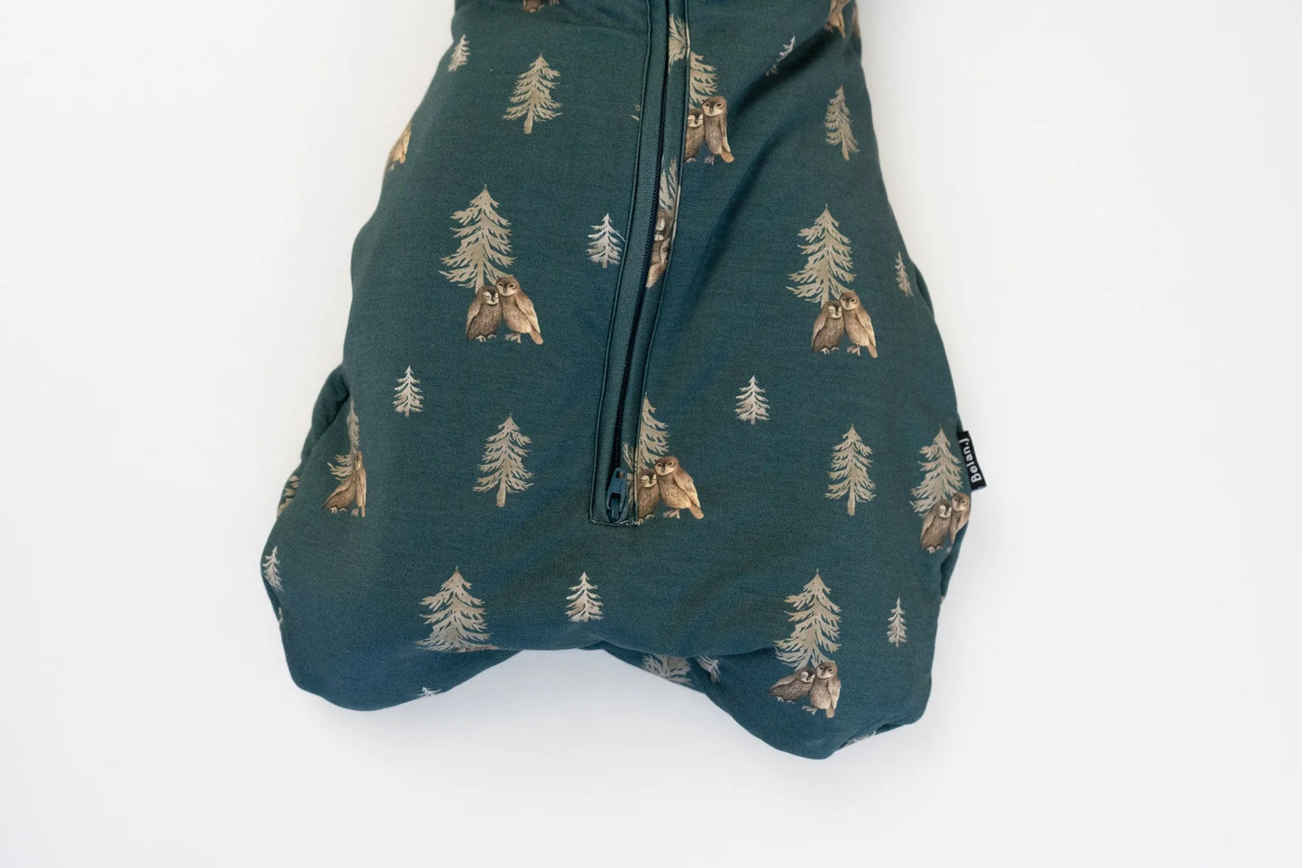 
                  
                    A close-up of a dark teal fabric item featuring a repeating pattern of owls and pine trees, with a central zipper, shown against a plain white background.
                  
                