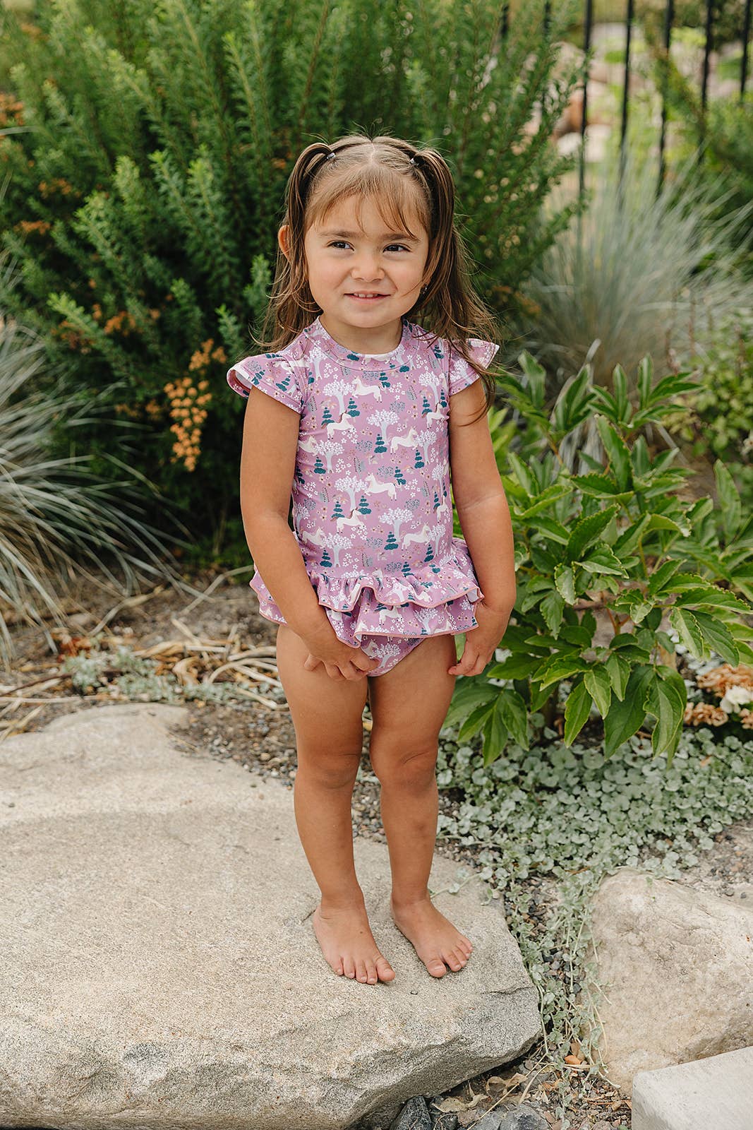 Child in a pink patterned swim outfit standing on rocks with greenery in the background