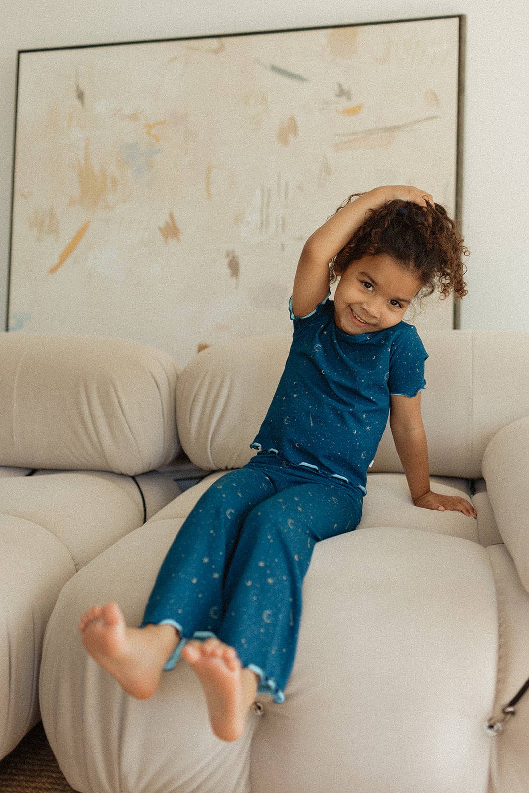 Child in a blue outfit sitting on a beige sofa with a colorful abstract painting in the background.