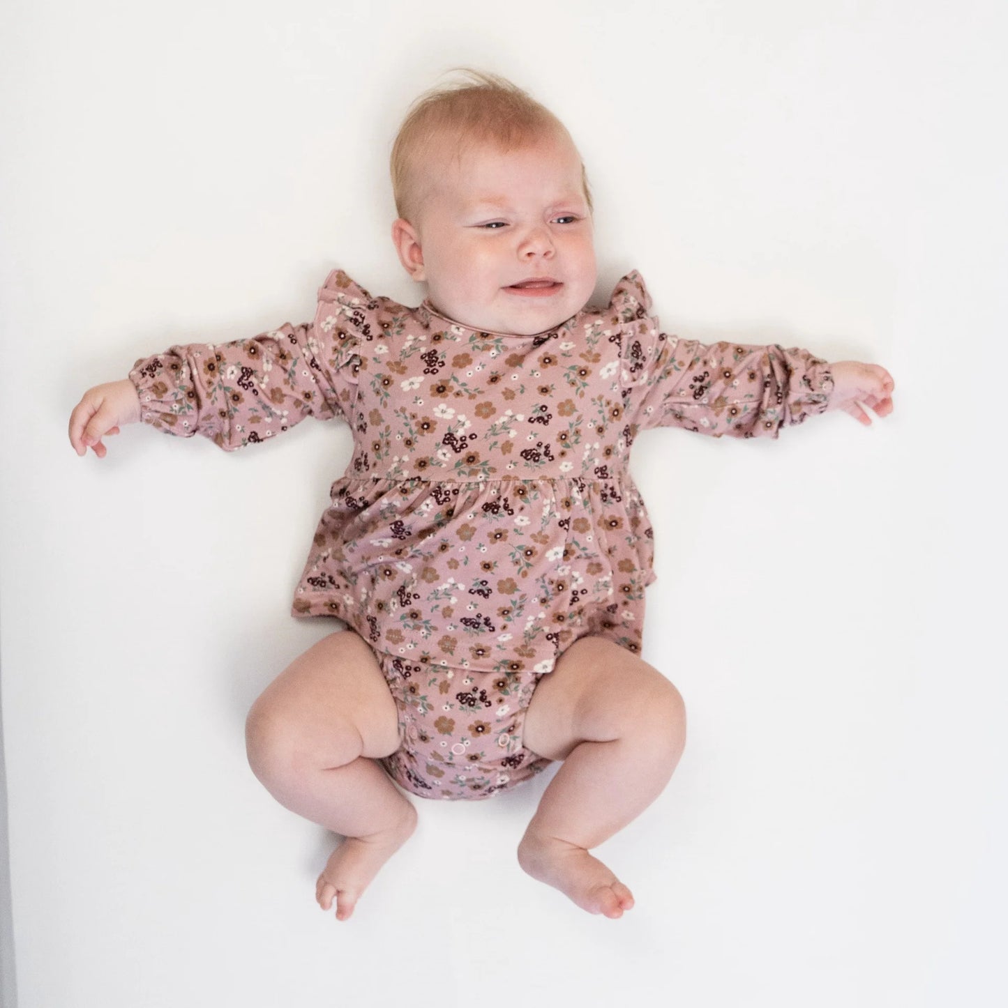 
                  
                    A baby lying on their back wearing a dusty pink long-sleeve romper with a brown and black floral pattern, set against a plain white background.
                  
                
