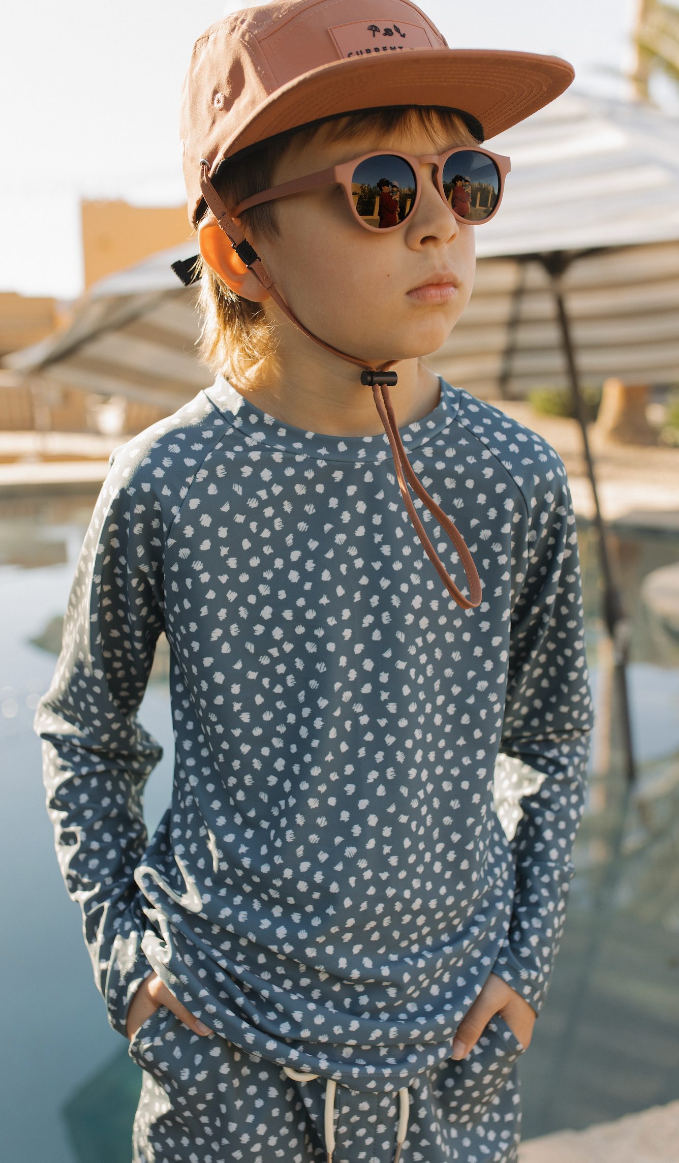 Child wearing a patterned shirt and sunglasses with a blurred outdoor background.