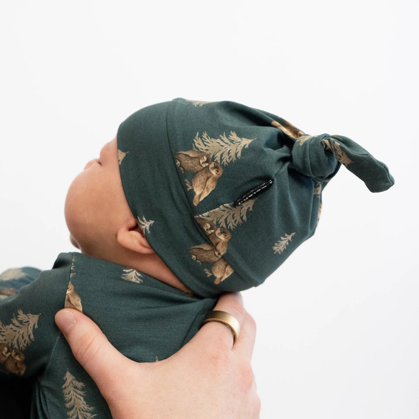 
                  
                    A baby wearing a dark green knotted beanie hat and matching swaddle blanket with a brown forest tree print, held by an adult hand against a plain white background.
                  
                
