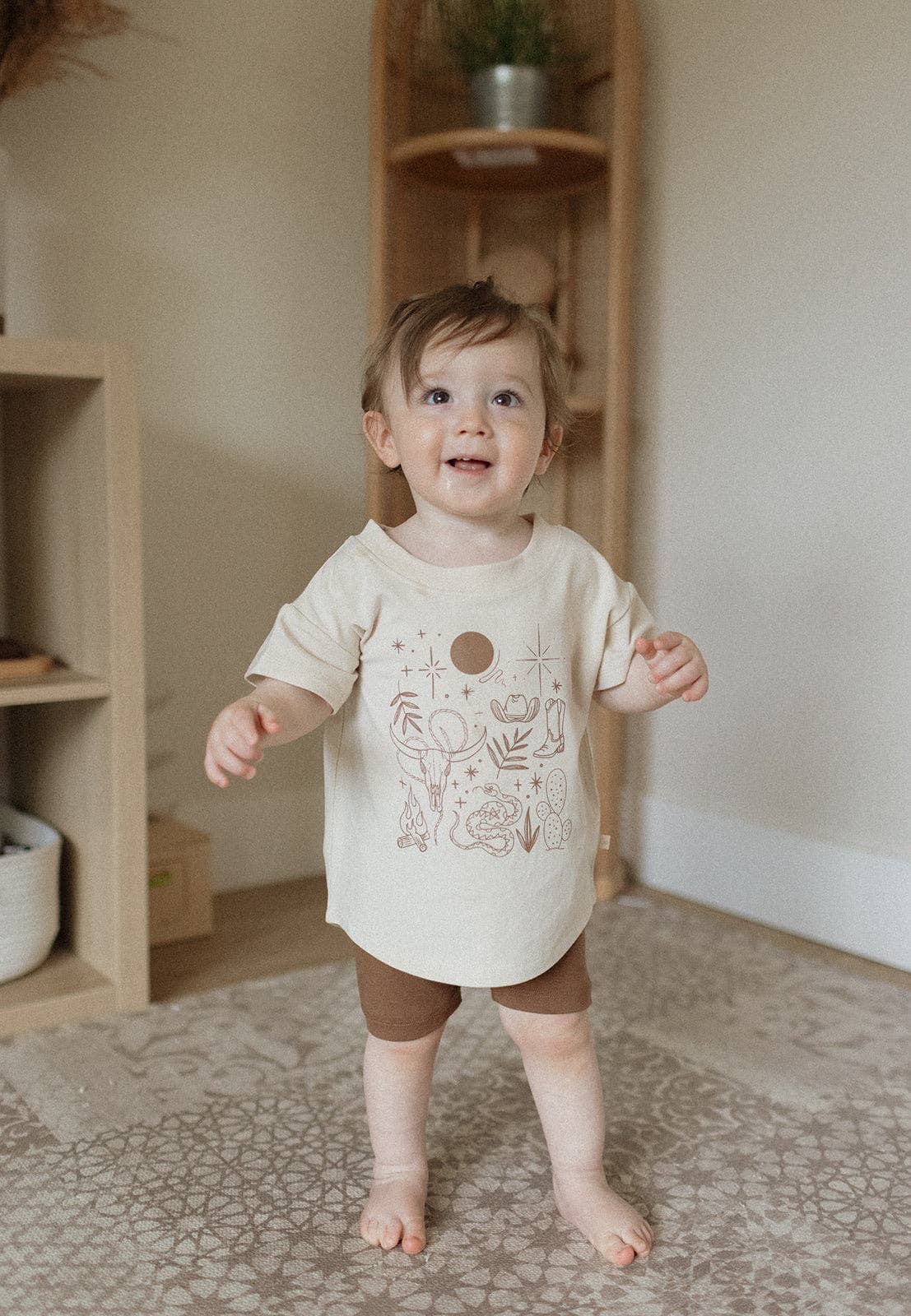 
                  
                    A toddler standing barefoot indoors, wearing a cream short-sleeve tunic top with a brown celestial graphic and matching brown shorts.
                  
                