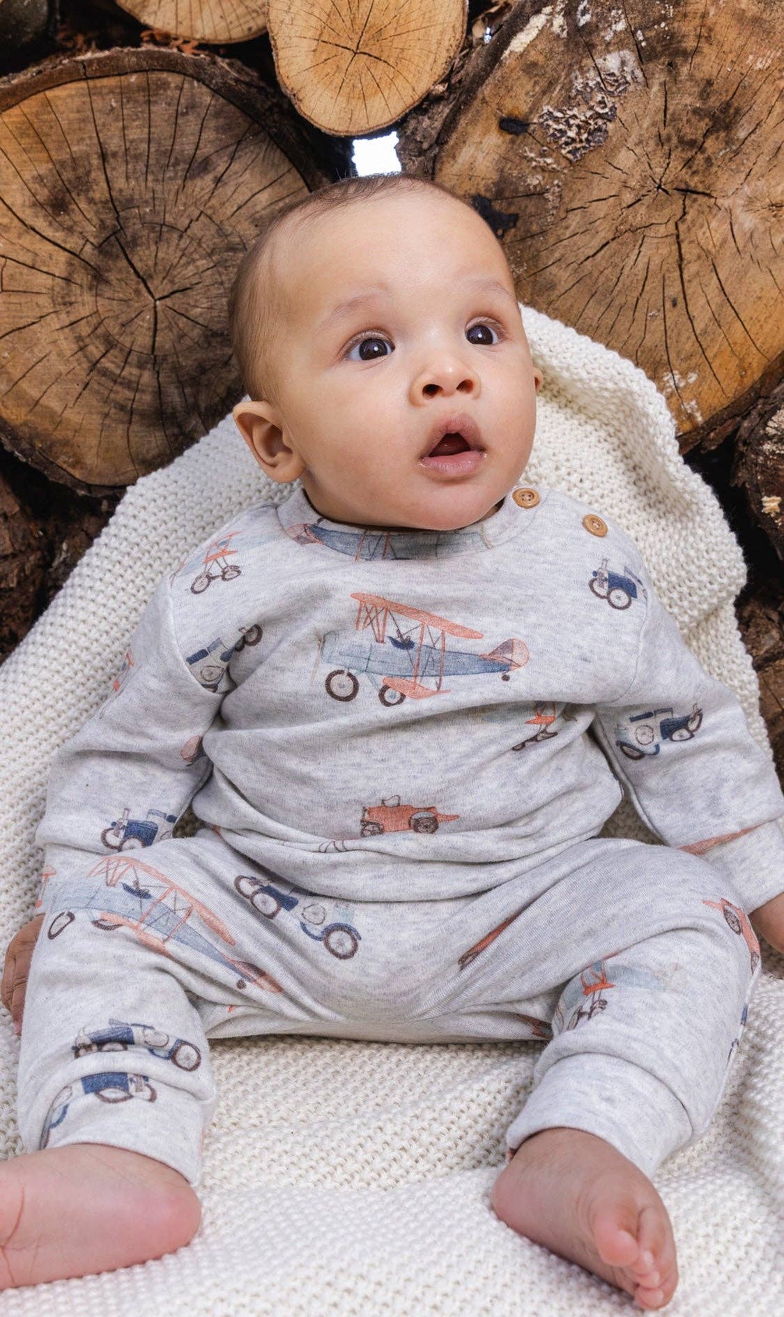 
                  
                    Baby in an airplane patterned outfit sitting on a wooden surface.
                  
                