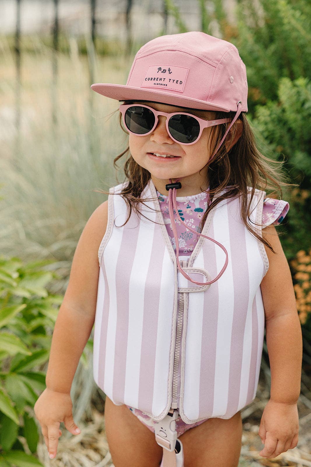 
                  
                    Child wearing a pink cap, sunglasses, and a striped outfit outdoors.
                  
                