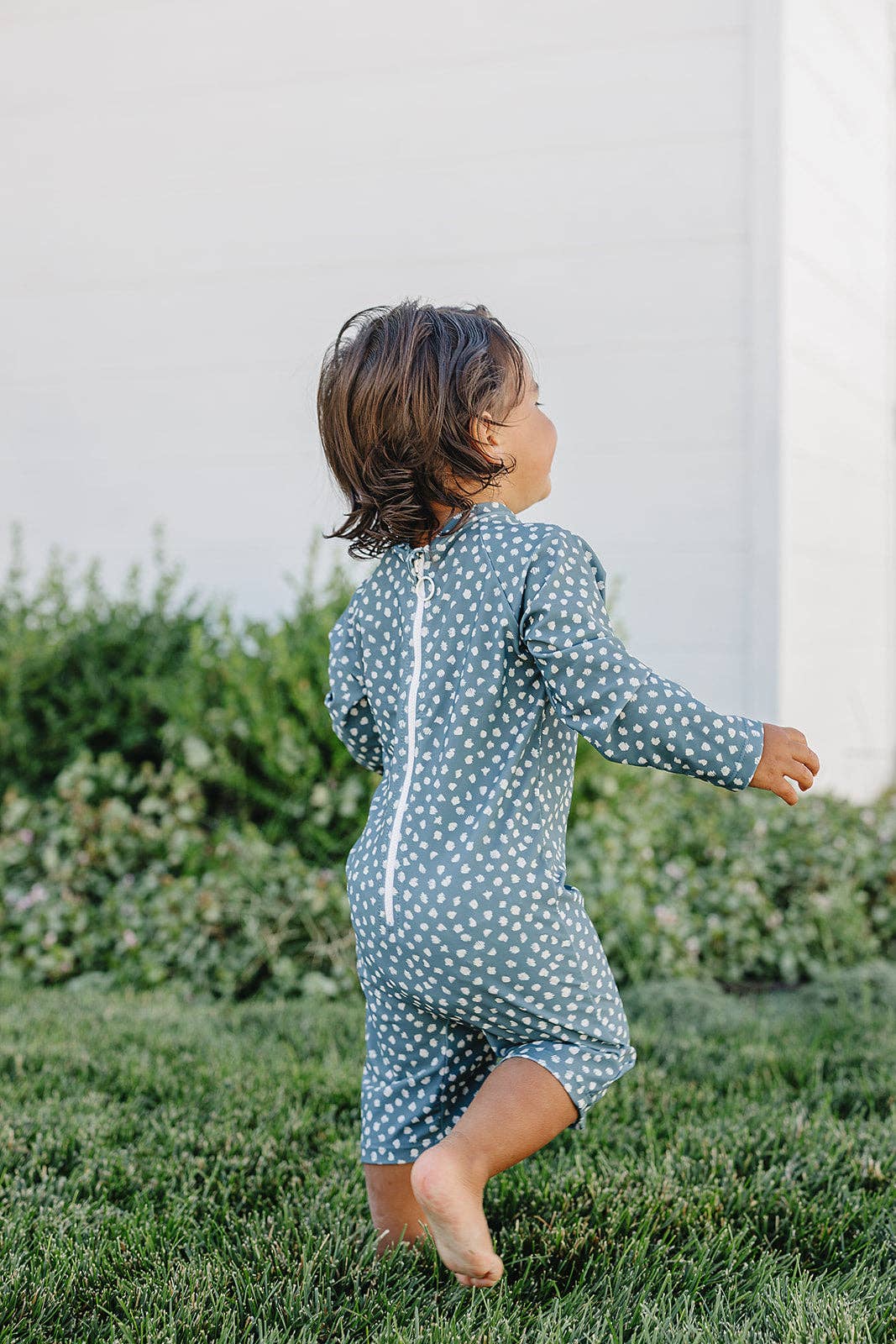
                  
                    Child wearing a blue polka dot sunsuit running on grass with a white wall and plants in the background
                  
                