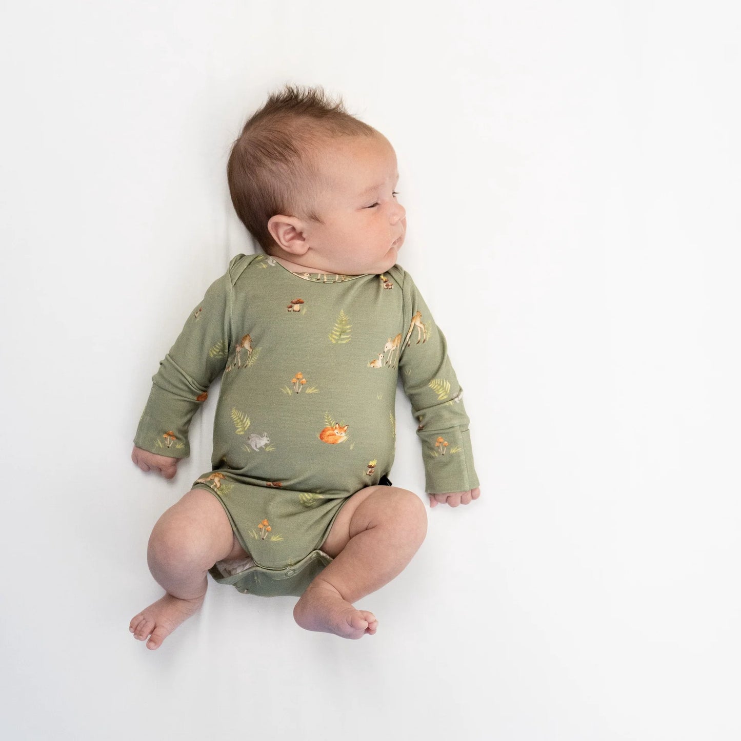 
                  
                    A baby lying on their side wearing a sage green long-sleeve bodysuit with a small woodland animal and flower print, photographed against a clean white background.
                  
                