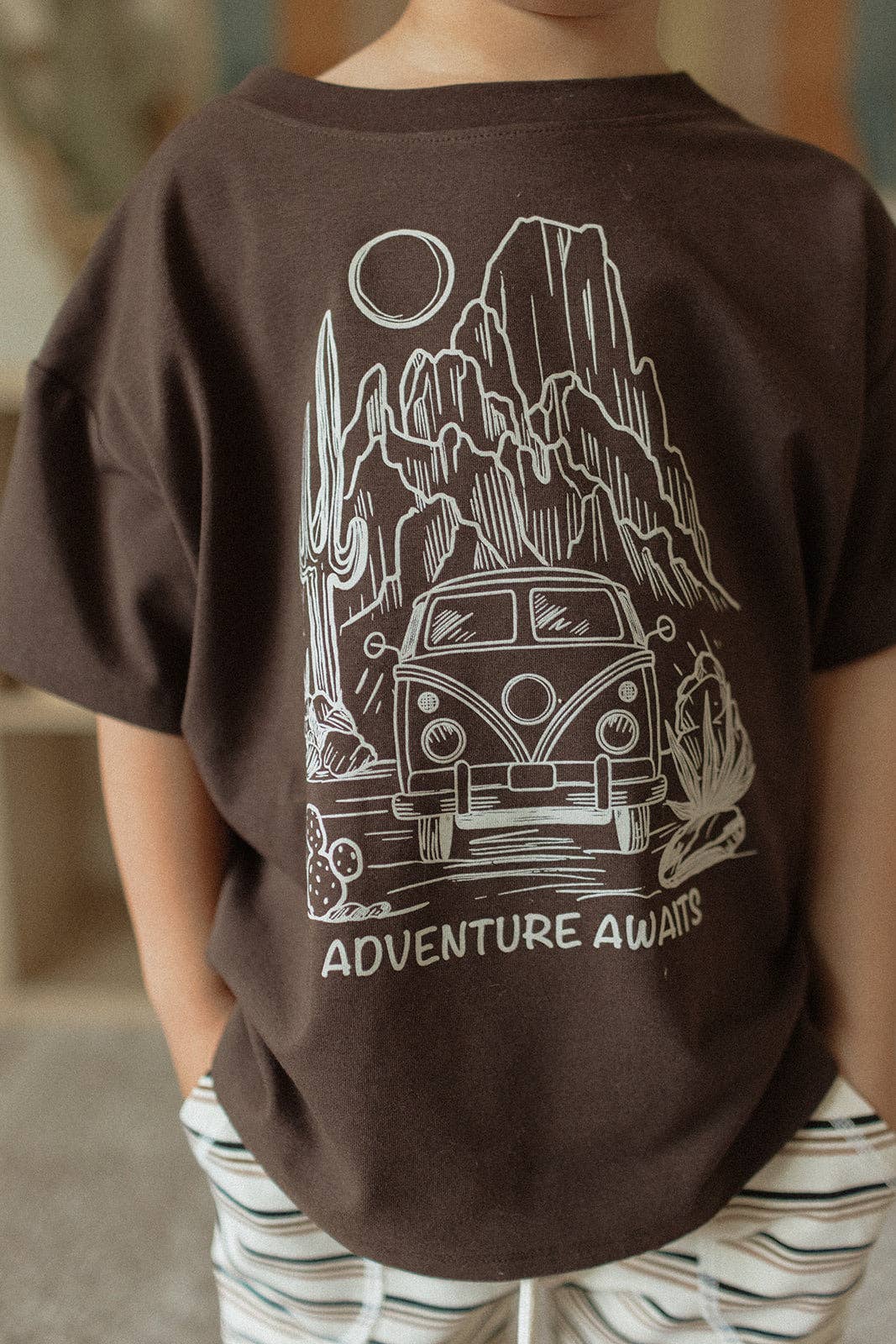 
                  
                    Child wearing a brown t-shirt with a graphic design and 'Adventure Awaits' text.
                  
                