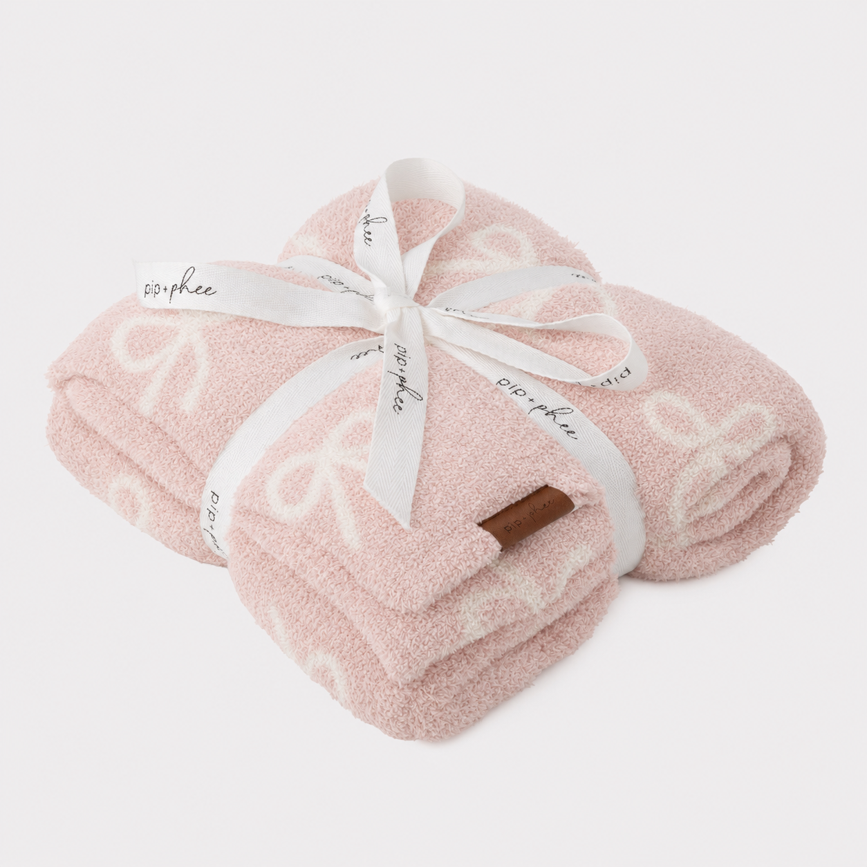 A folded light pink plush blanket with a white embossed bow pattern, wrapped in a white ribbon printed with the brand name 'pip + phee' and tied in a bow, on a light gray background.
