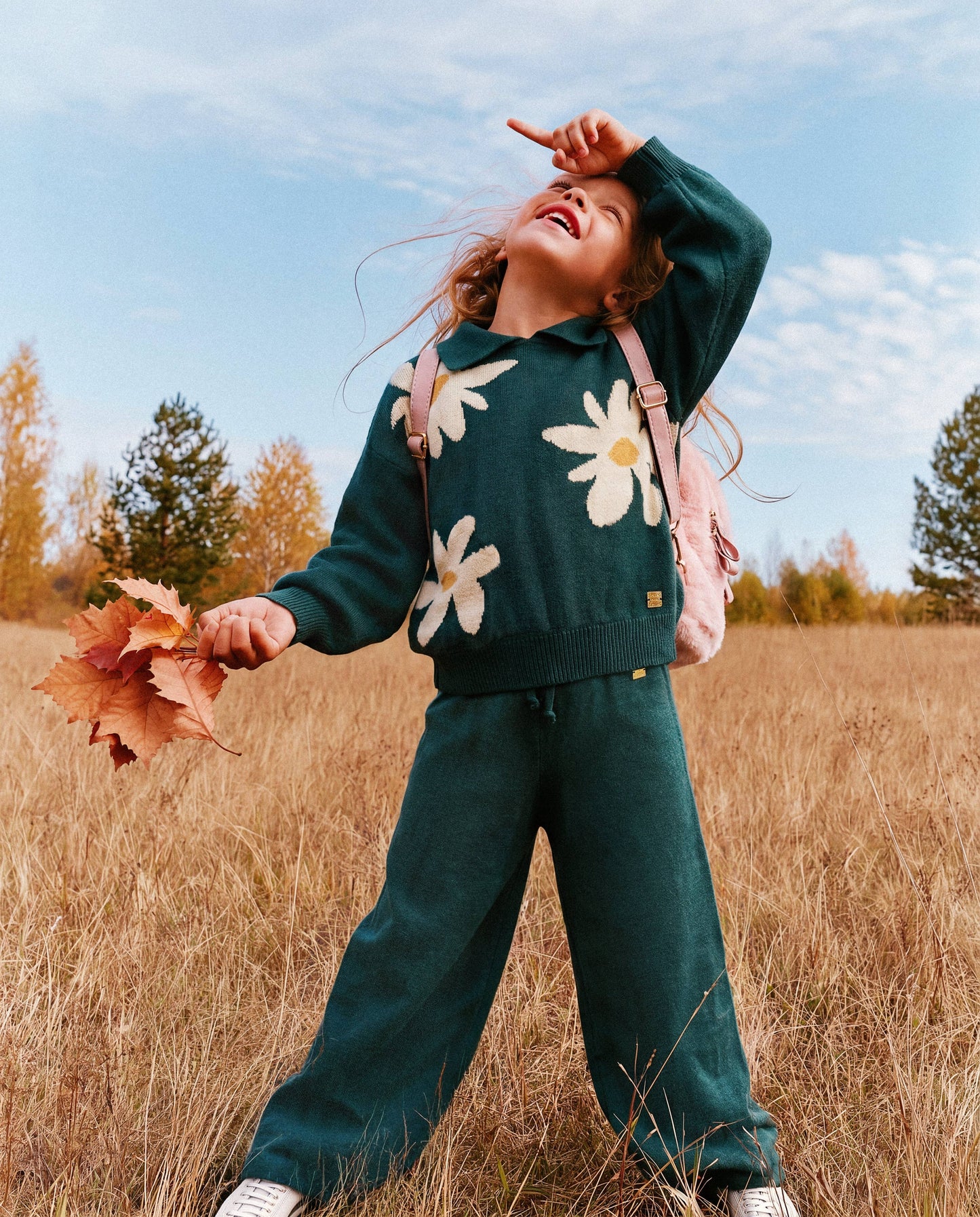 
                  
                    Child in a green floral sweater and pants standing in a field with a pink backpack, holding a leaf.
                  
                