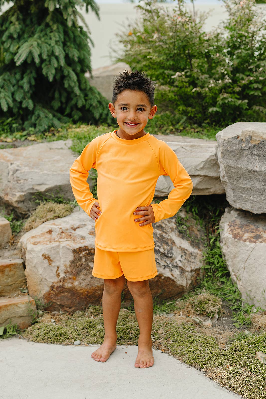 Child wearing an orange swim outfit standing outdoors near rocks and greenery.