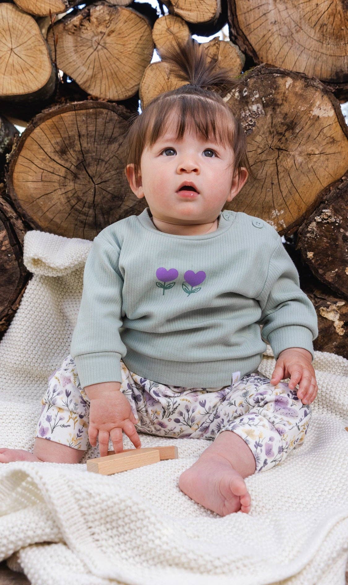
                  
                    Baby sitting on a blanket with wooden logs in the background.
                  
                