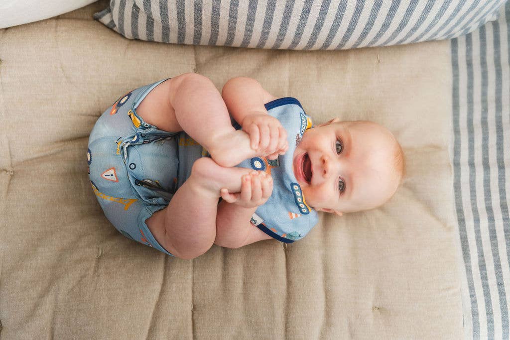 
                  
                    Baby lying on a beige blanket wearing a blue bib and outfit with cartoon designs.
                  
                