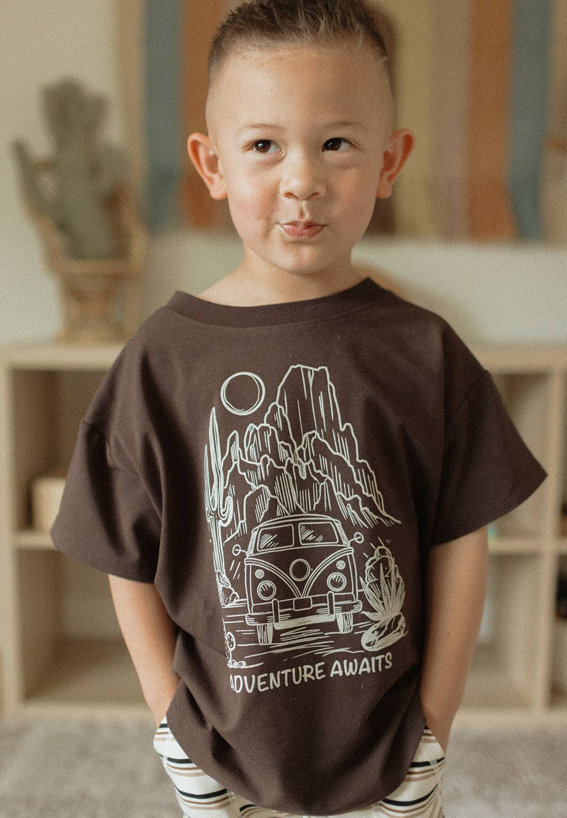 
                  
                    Child wearing a brown t-shirt with a graphic design and text, standing indoors.
                  
                