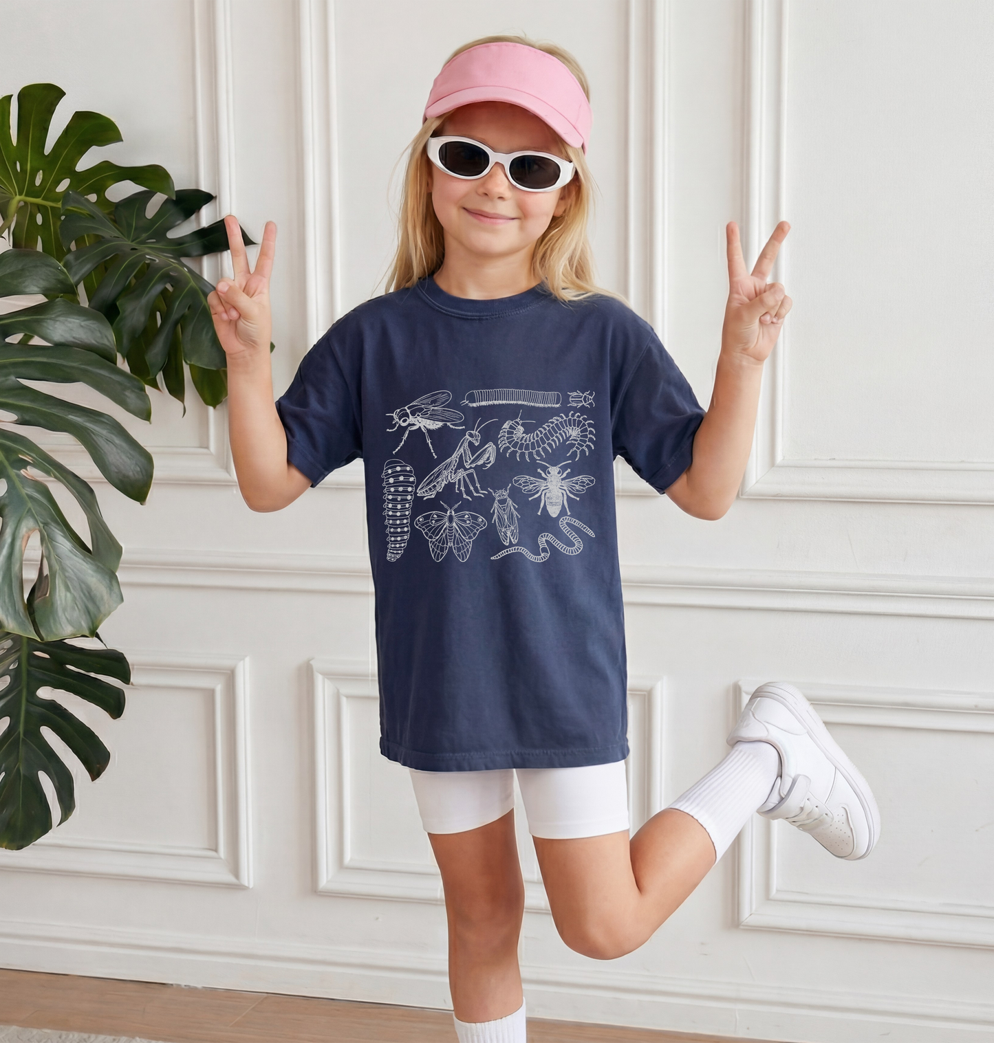 Child wearing a navy blue t-shirt with insect illustrations, white shorts, and a pink visor, posing indoors.