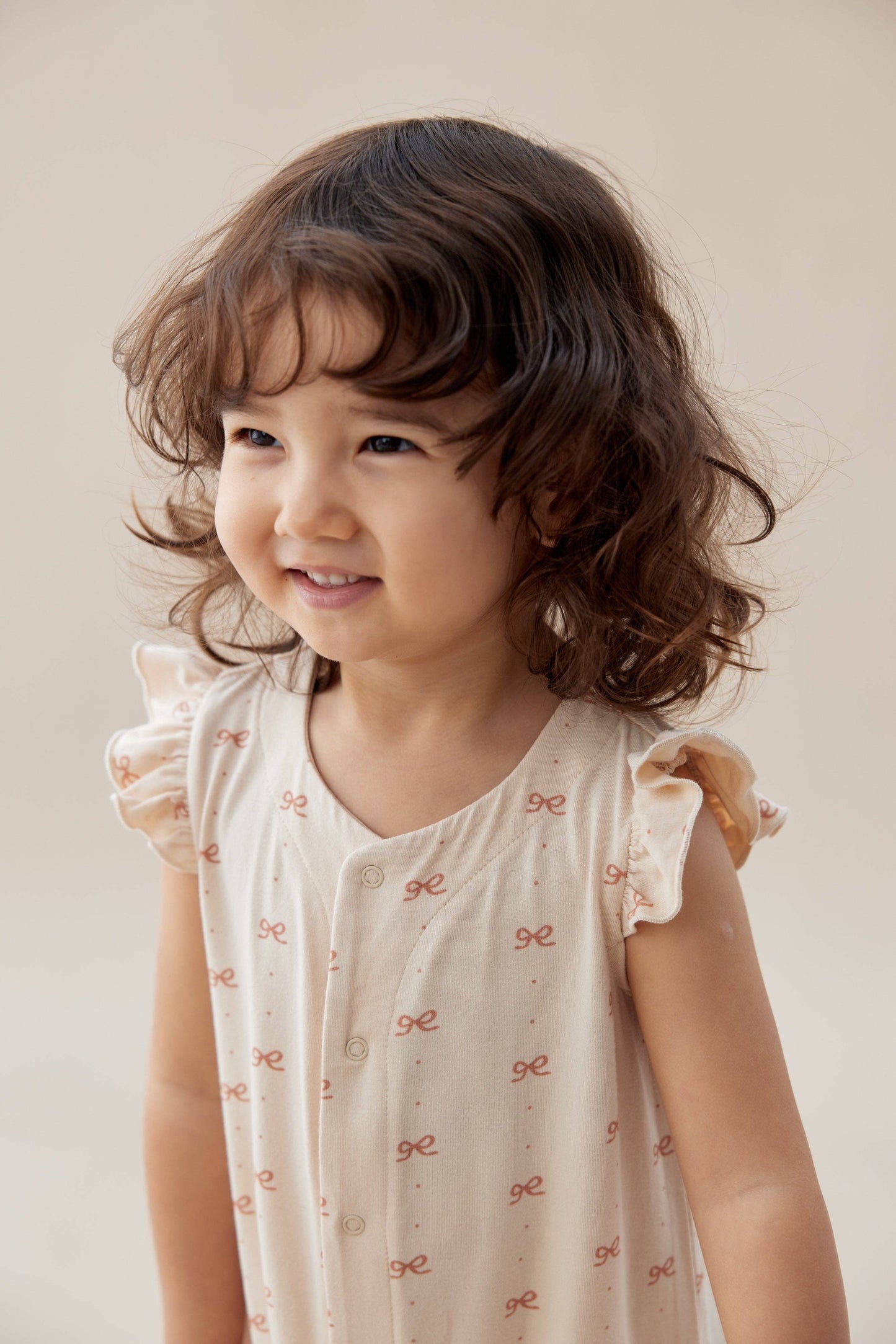 
                  
                    Young girl wearing a light-colored romper with ruffled sleeves against a plain background.
                  
                