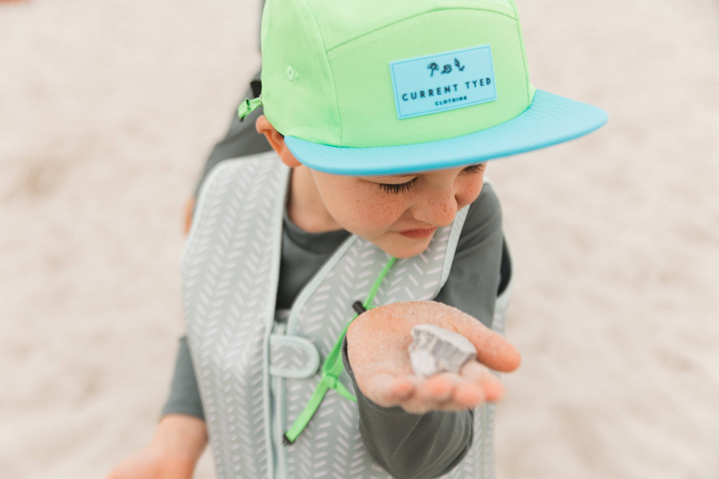 
                  
                    Child wearing a green and blue cap with a brand logo, holding sand on a beach.
                  
                