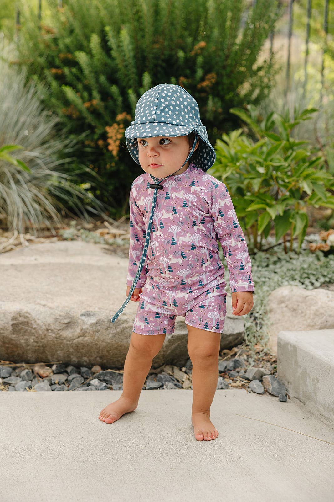 
                  
                    Child wearing a patterned sun hat and unicorn patterned swimsuit standing outdoors.
                  
                