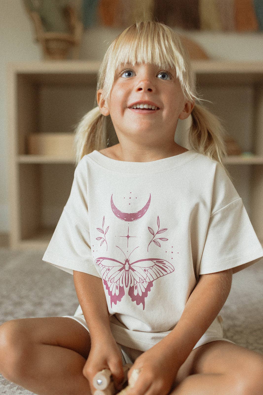 Child wearing a white t-shirt with pink butterfly design sitting in a room with wooden shelves.