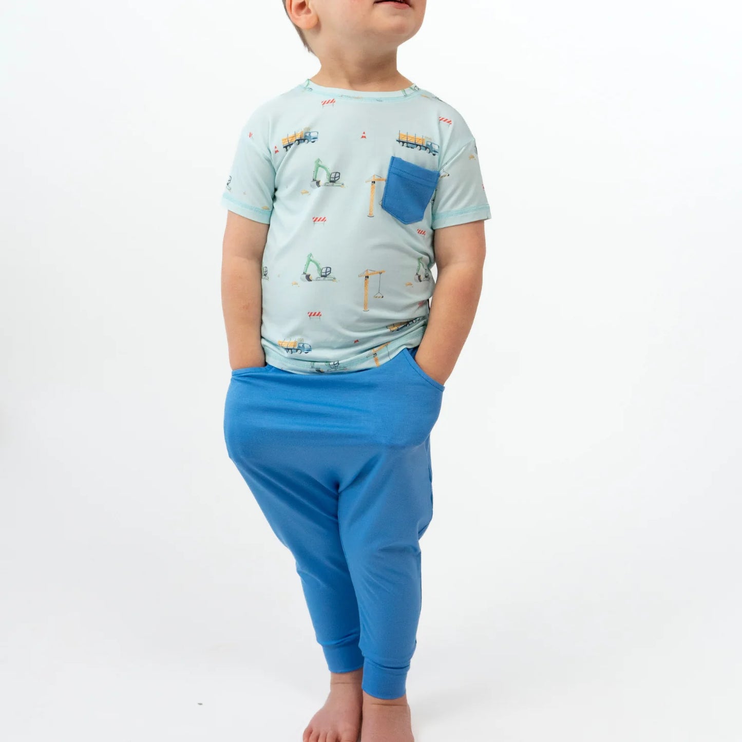 
                  
                    Child wearing a light blue shirt with vehicle prints and blue pants on a white background
                  
                