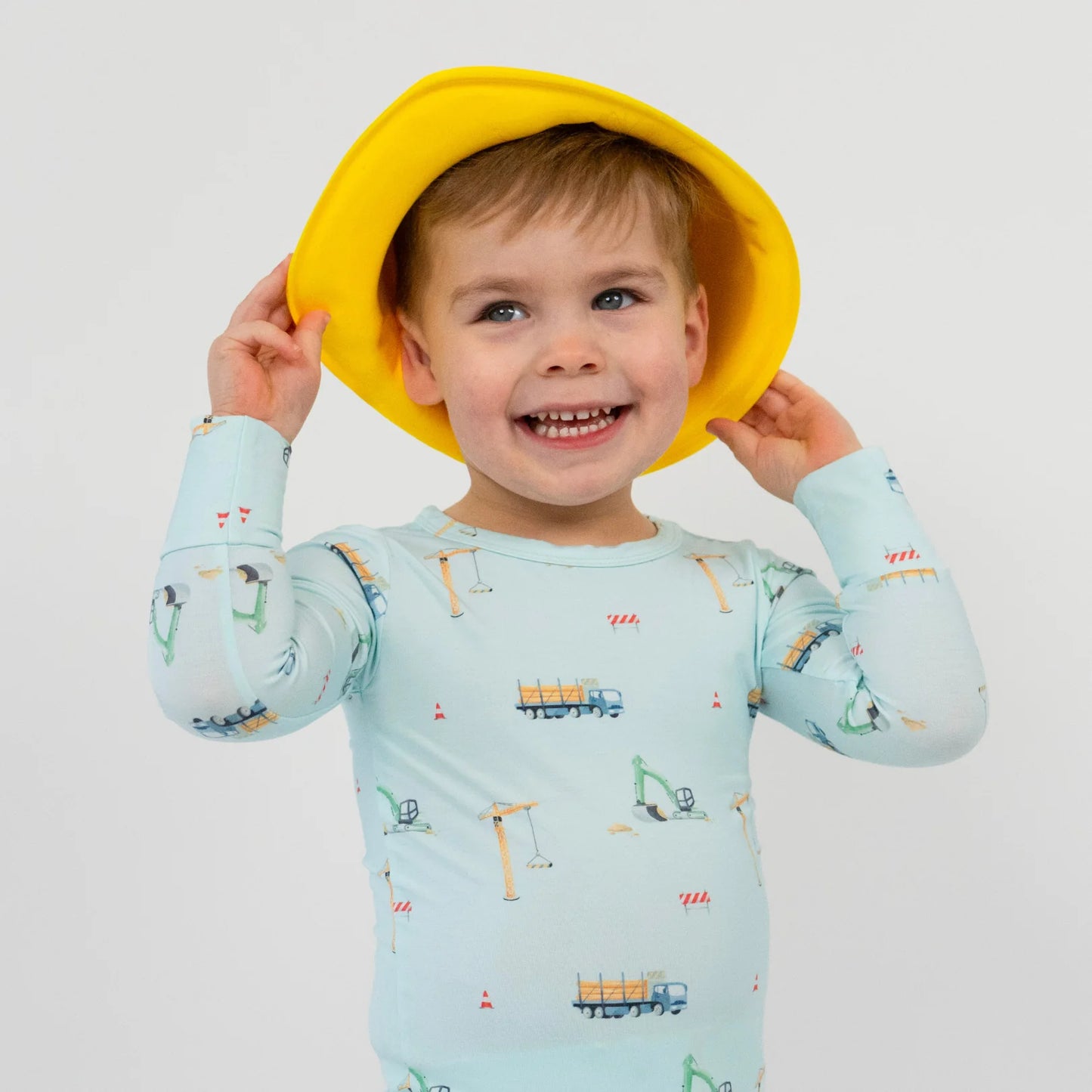
                  
                    Child wearing a light blue pajama shirt with a construction vehicle print and a yellow hat on a white background. 
                  
                