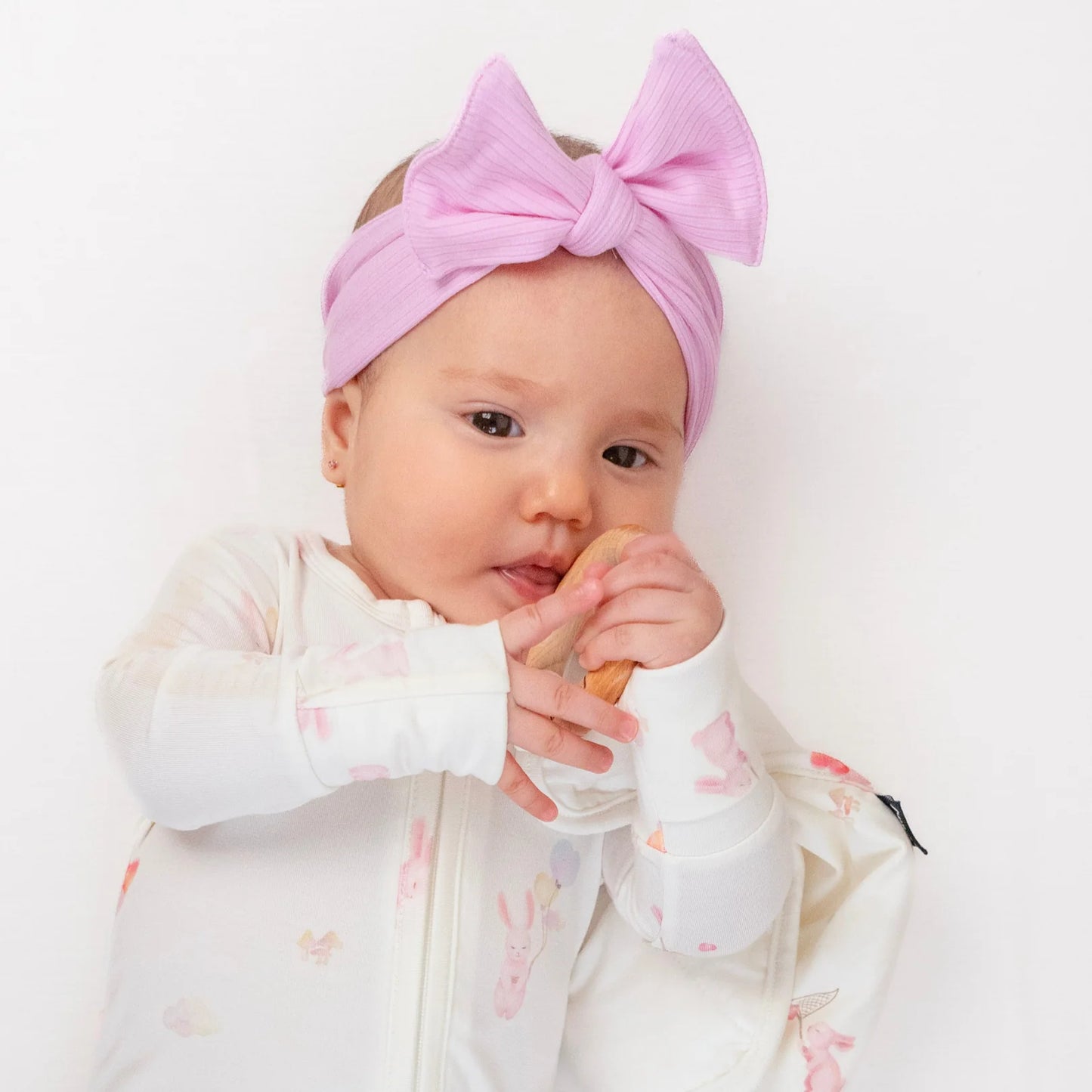 
                  
                    Baby wearing a pink headband with a bow, sitting on a white surface.
                  
                