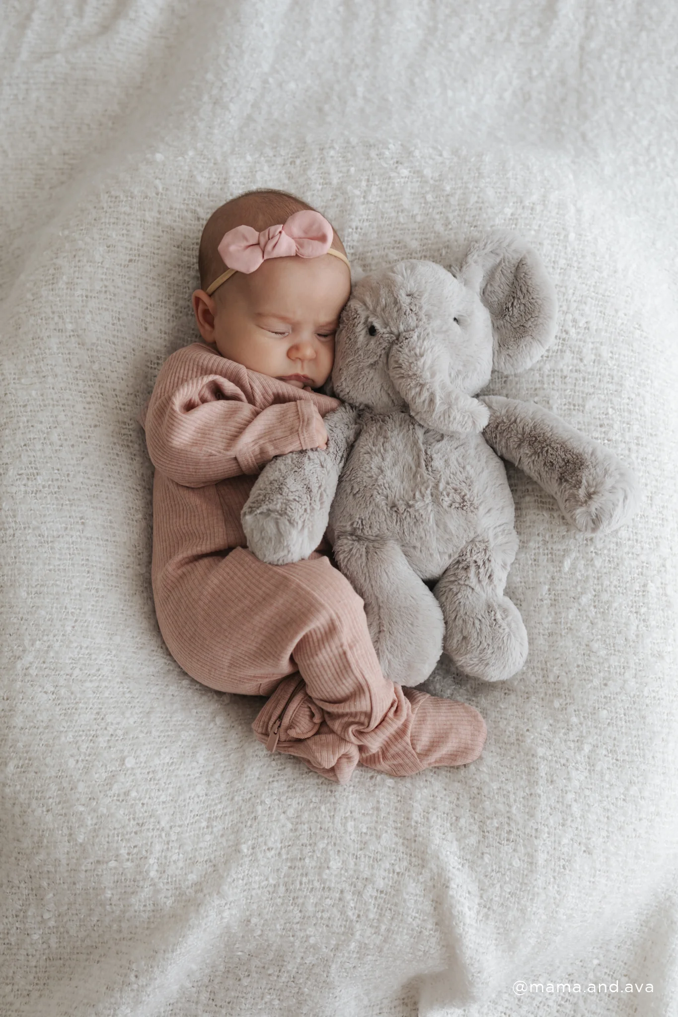 
                  
                    Newborn baby in pink outfit with a gray elephant plush toy on a soft surface.
                  
                