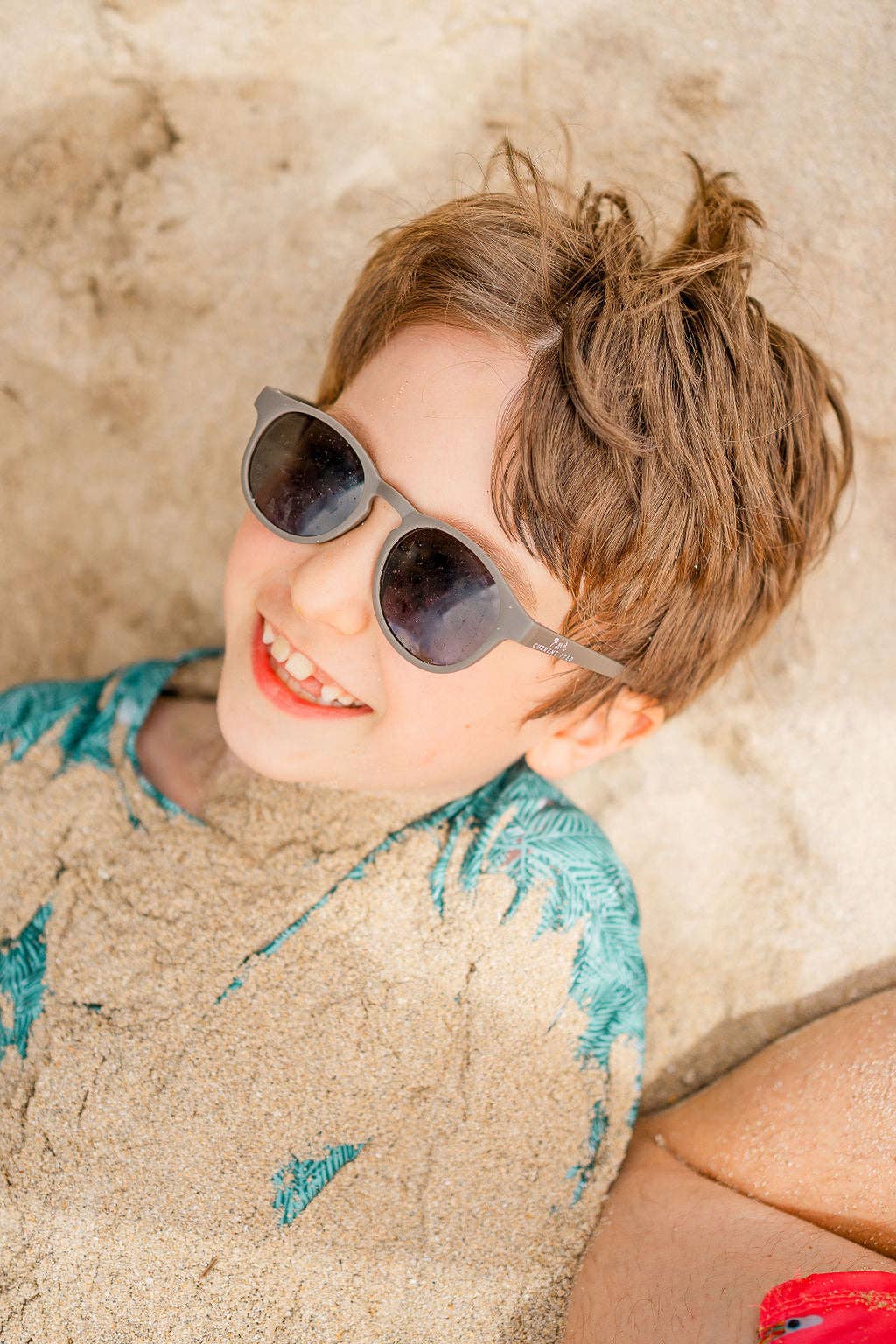 Child wearing sunglasses and a blue shirt lying on sand.