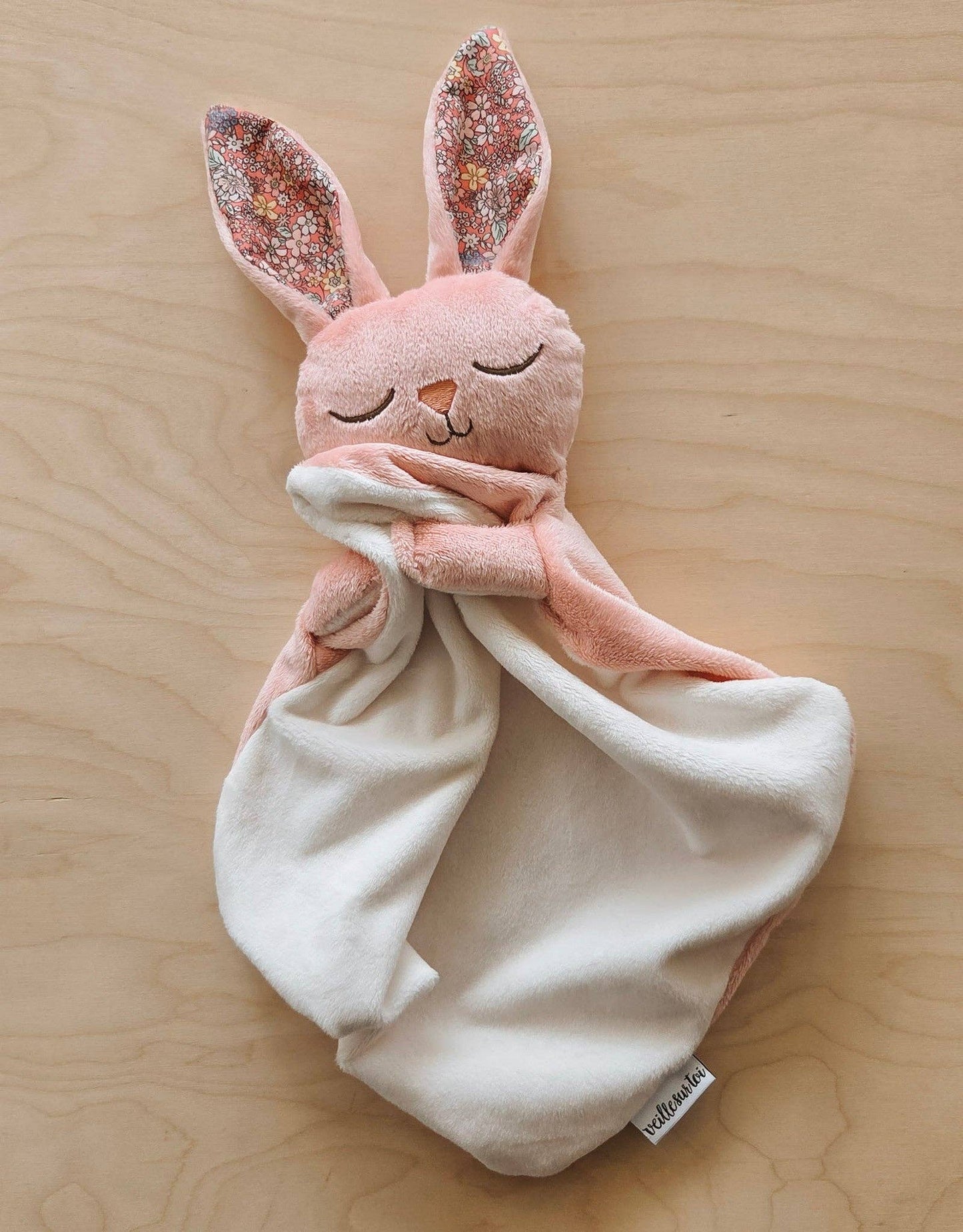
                  
                    Pink plush bunny toy with a white blanket on a wooden surface.
                  
                