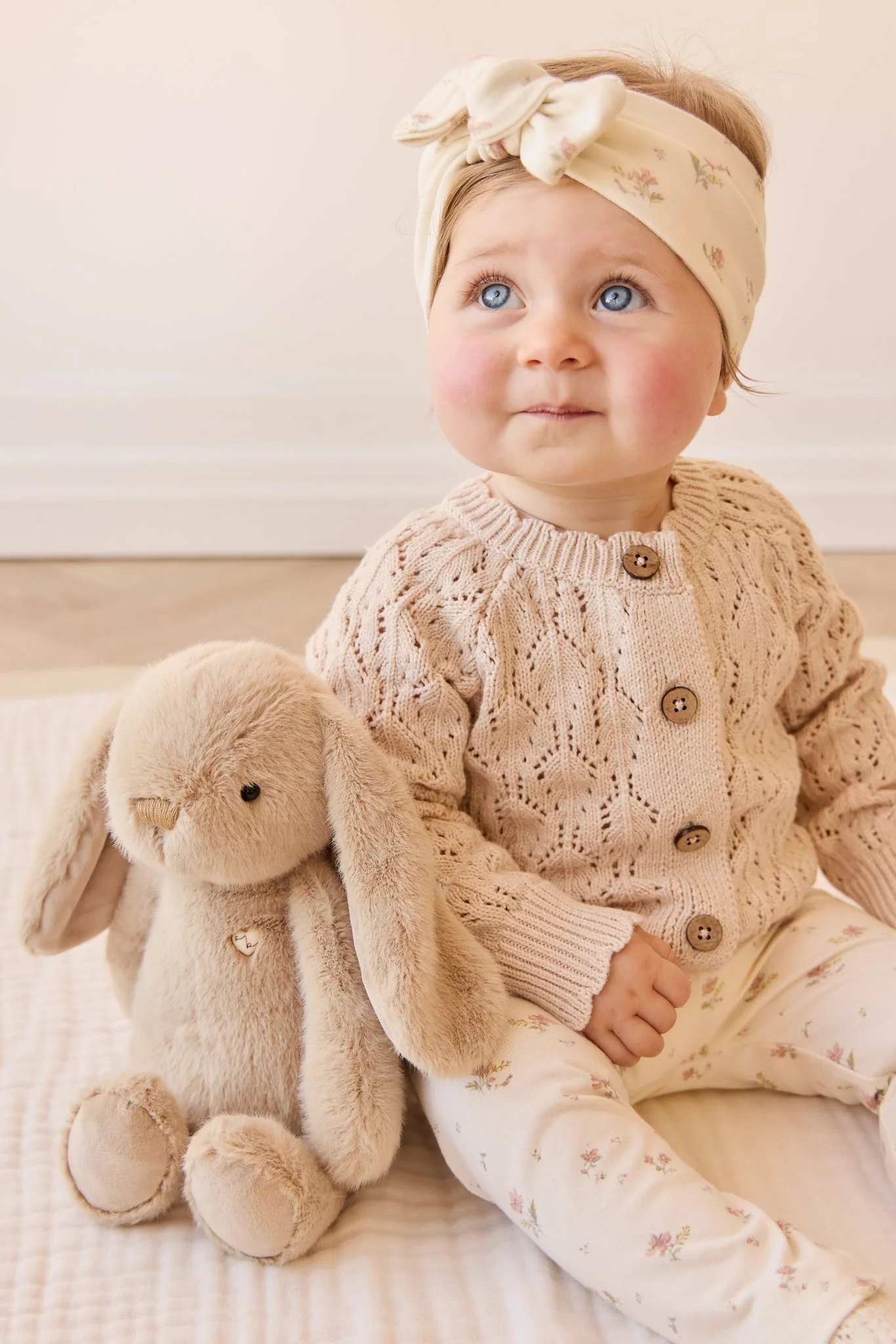 
                  
                    Baby in a knitted outfit sitting next to a plush toy on a soft surface.
                  
                