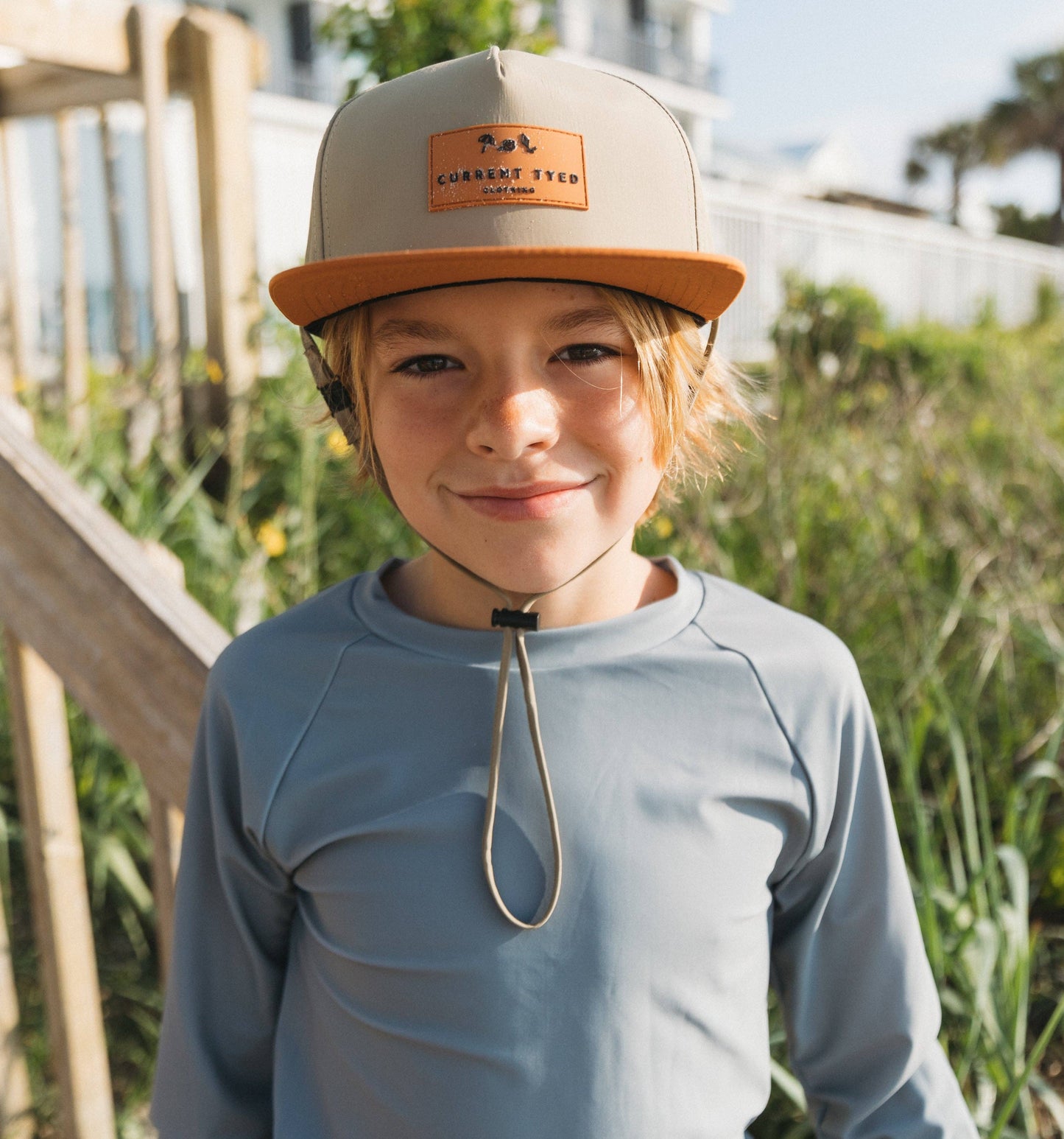 Child wearing a beige and brown cap with a logo, standing outdoors.