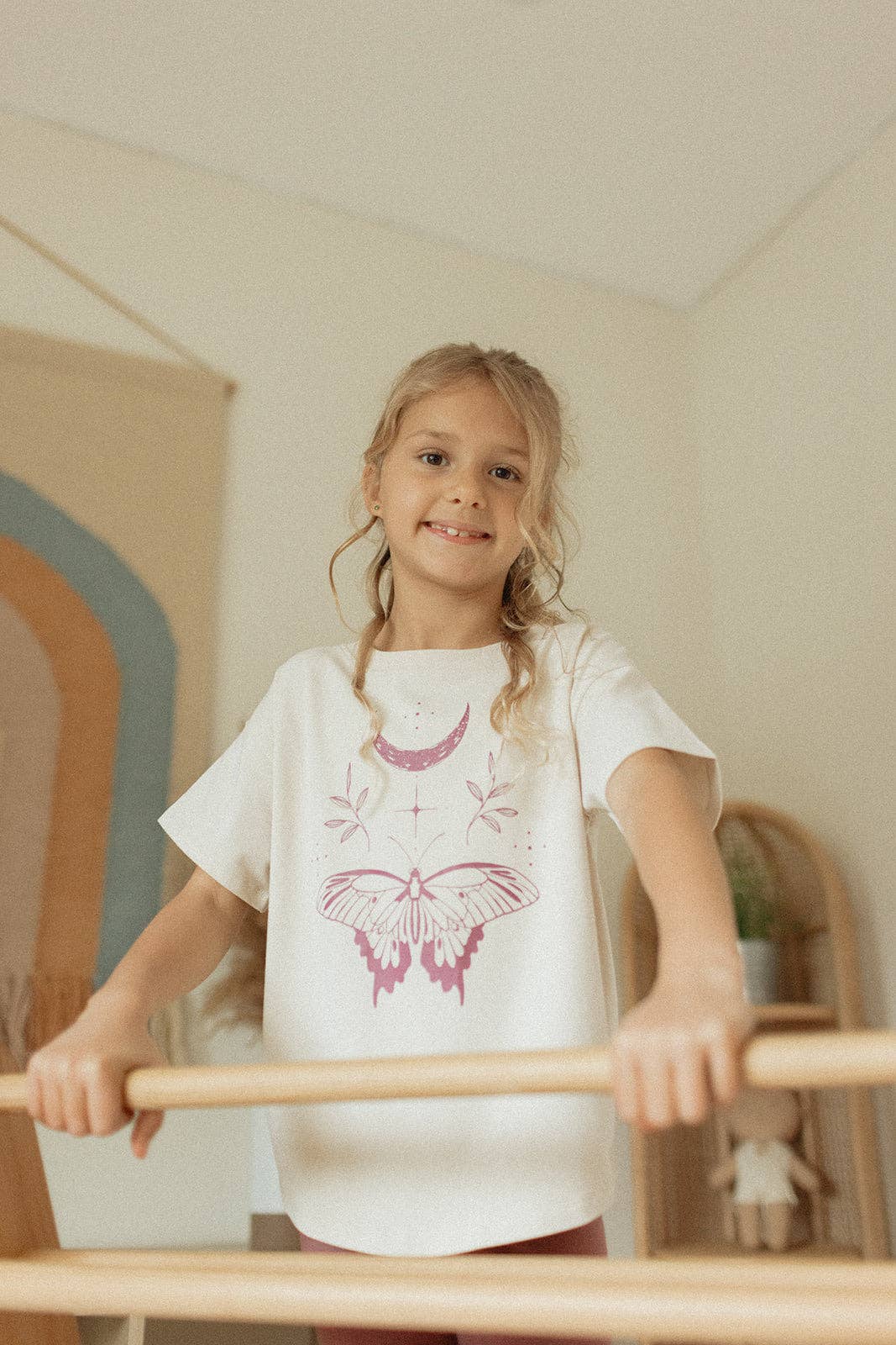 
                  
                    Young girl wearing a white t-shirt with pink butterfly design, standing indoors.
                  
                