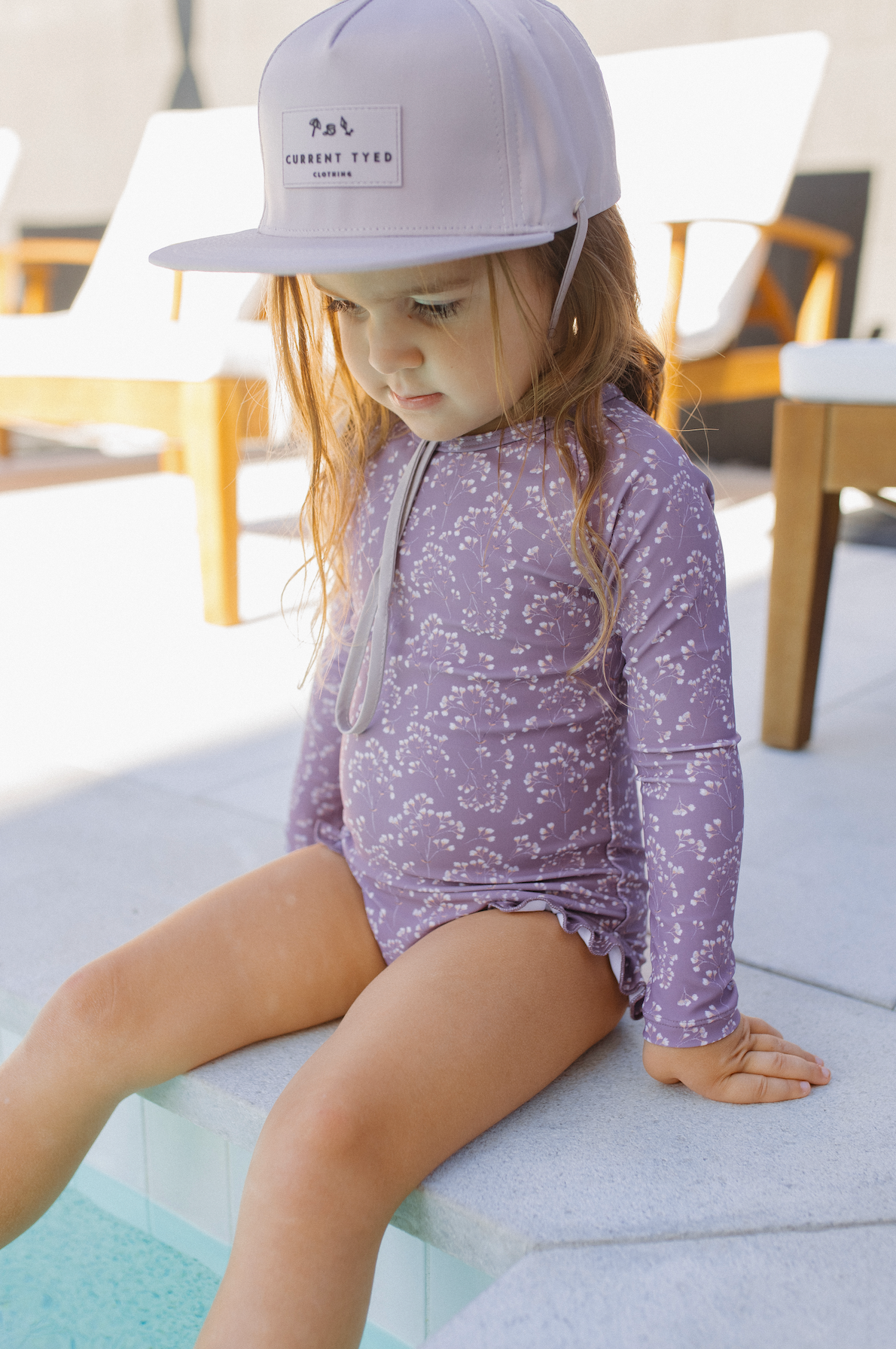 
                  
                    Child wearing a purple long-sleeve swimsuit and purple cap sitting on the edge of a pool. 
                  
                