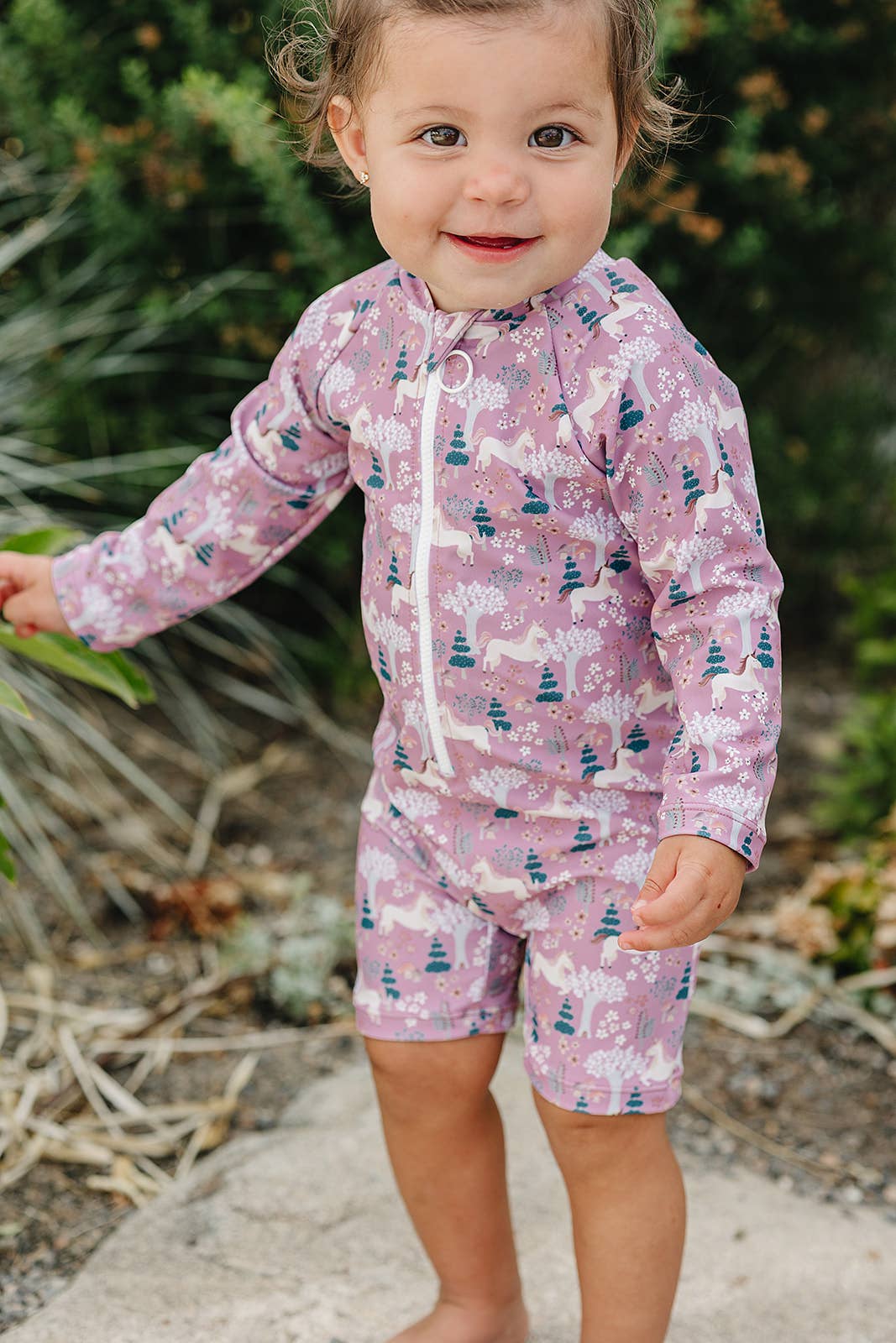 
                  
                    Child wearing a pink and purple unicorn patterned sunsuit outdoors.
                  
                