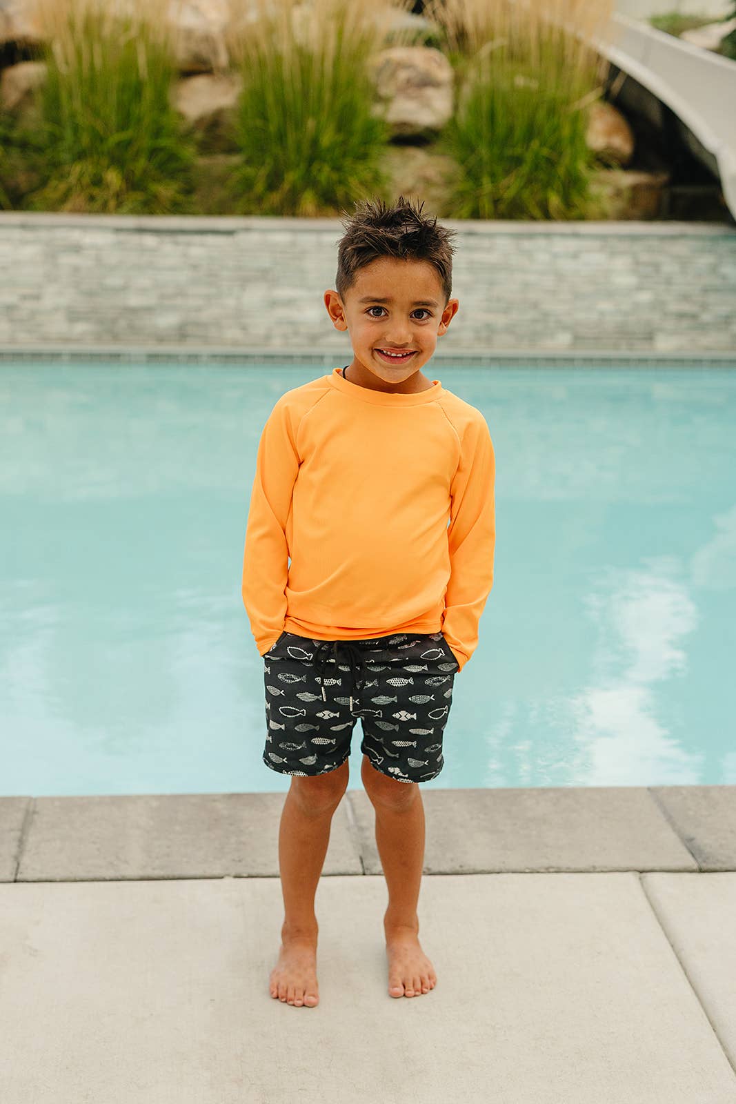 
                  
                    Child wearing an orange rash guard shirt and black swim shorts standing by a pool.
                  
                