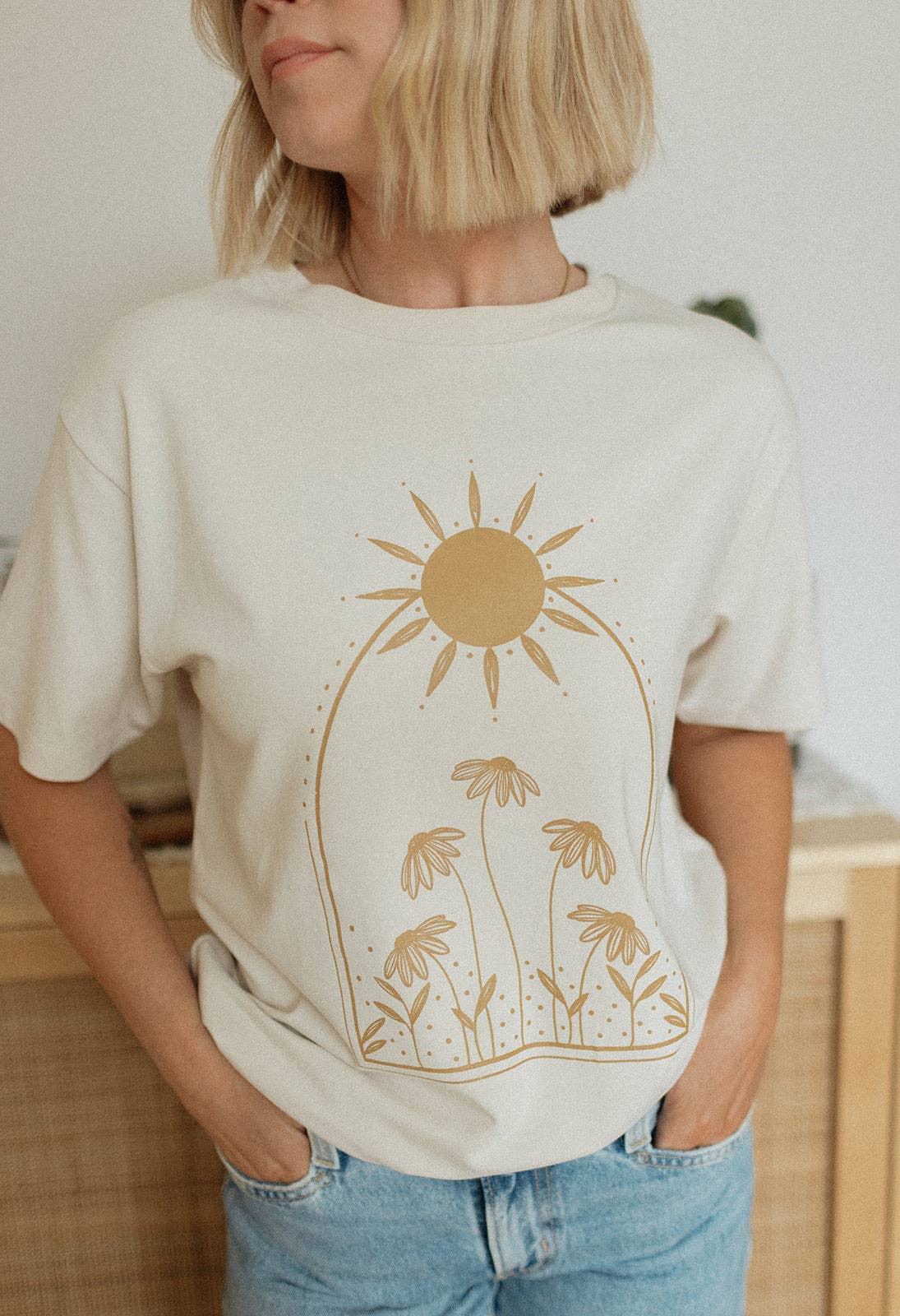 
                  
                    Person wearing a cream t-shirt with a sun and palm tree design, standing indoors.
                  
                