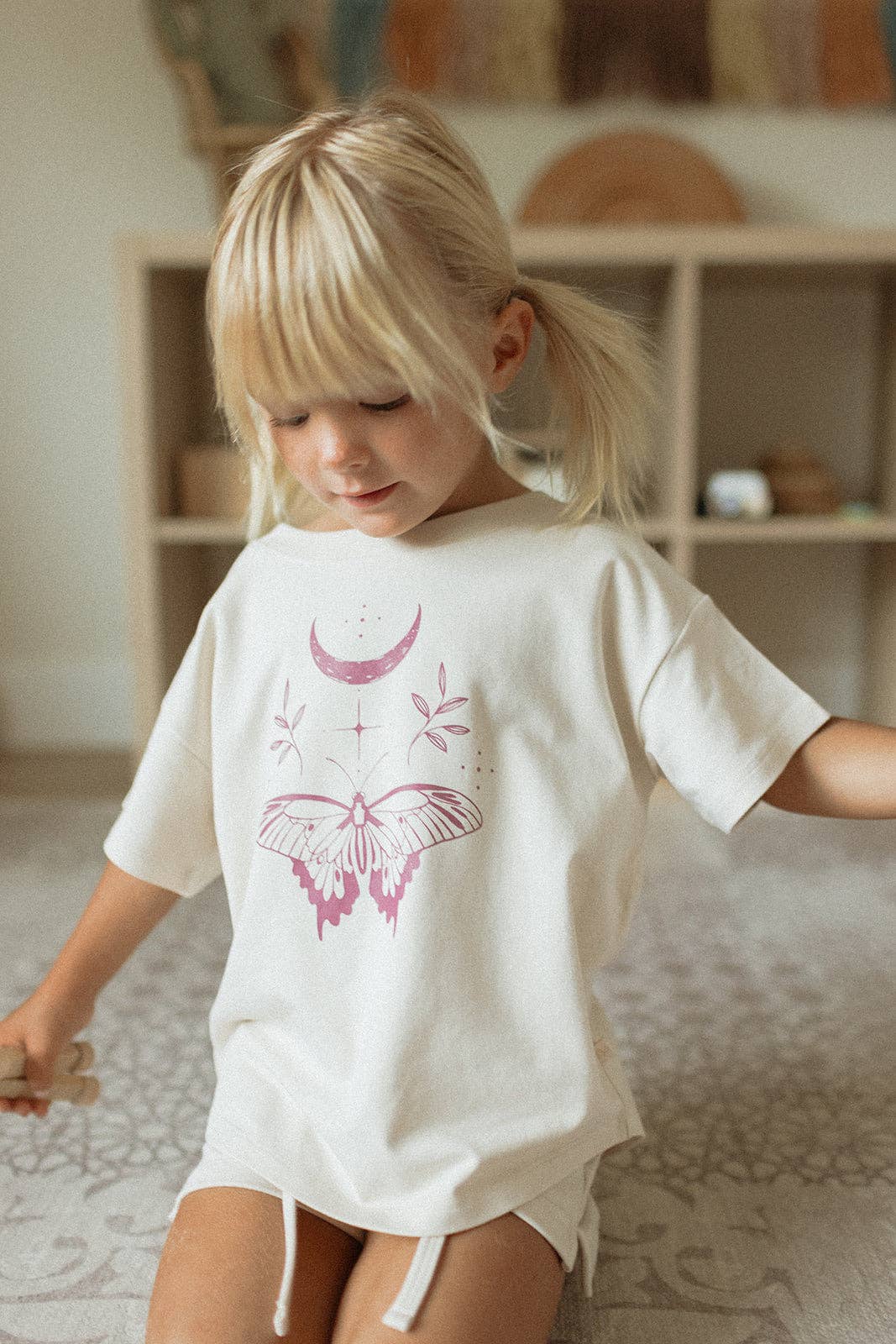 
                  
                    Child wearing a white t-shirt with pink butterfly design in a room with wooden shelves.
                  
                