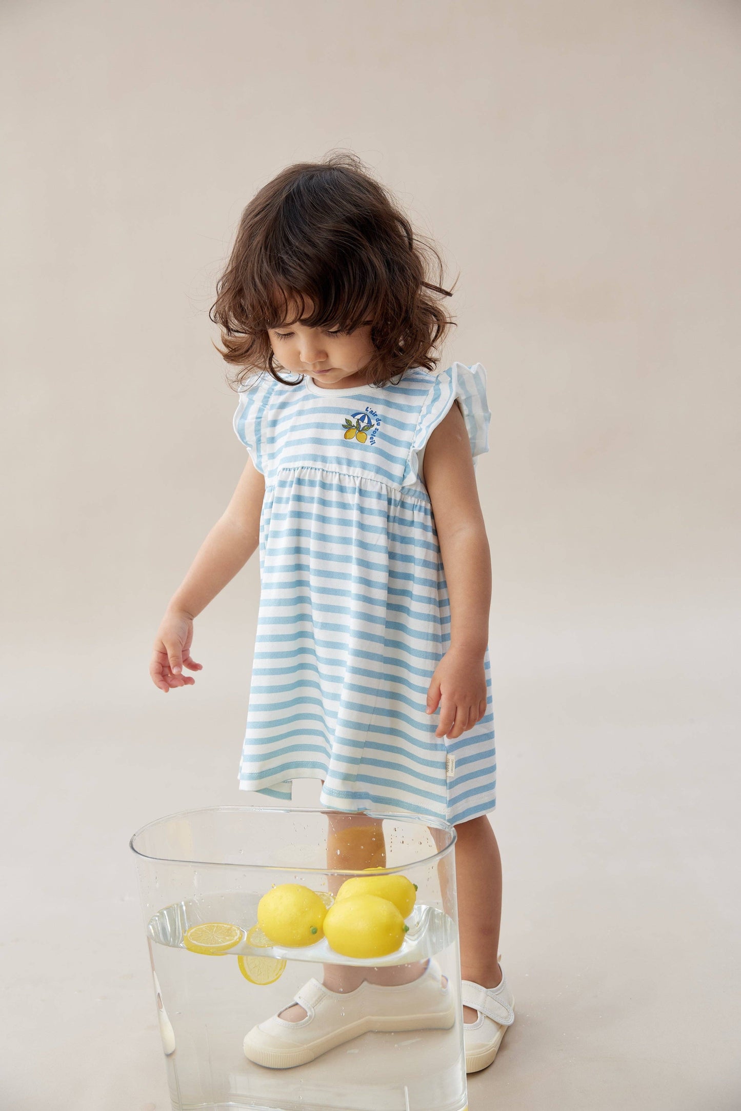 
                  
                    Child wearing a blue and white striped dress standing next to a glass table with lemons.
                  
                